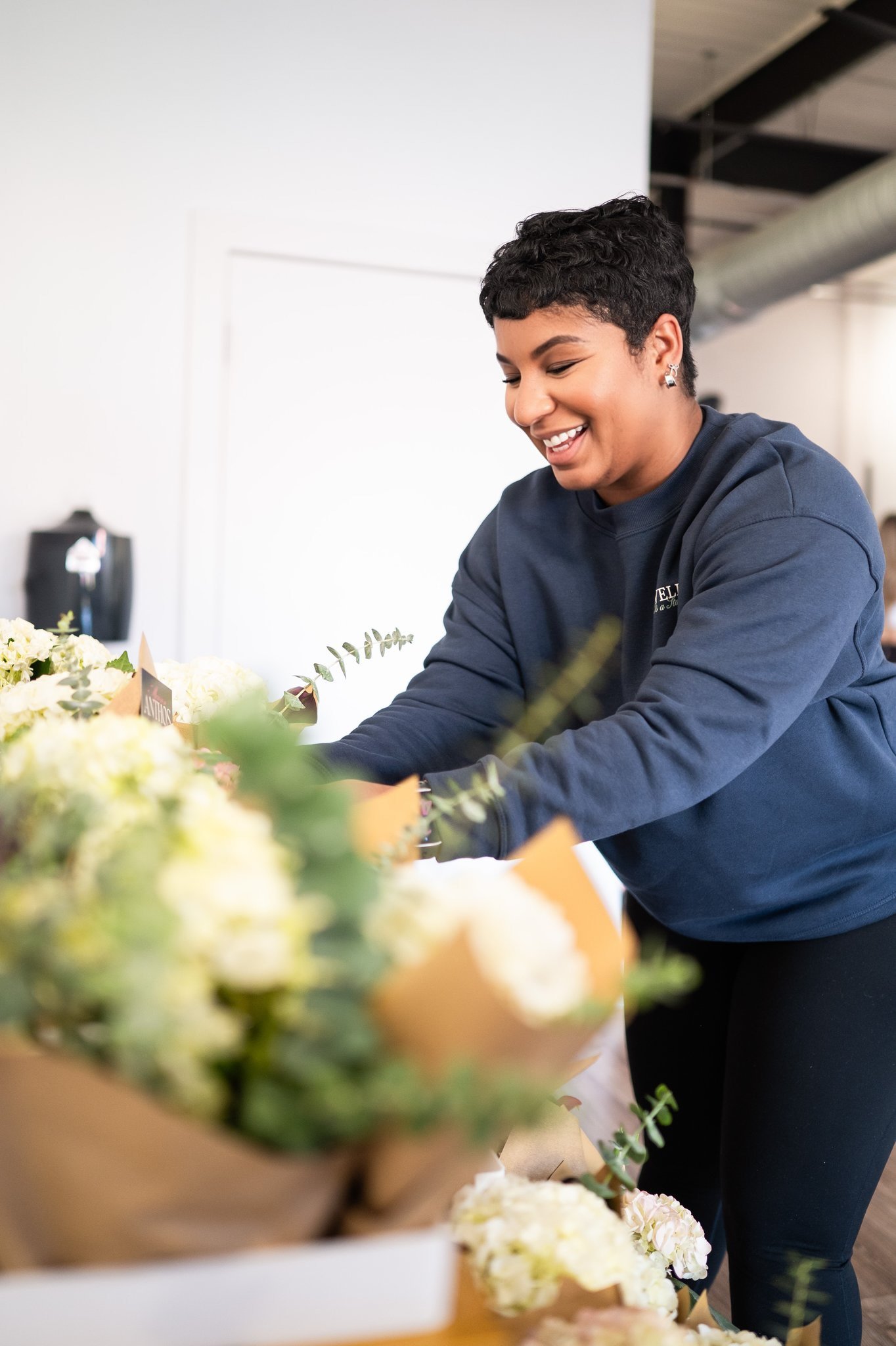 A young woman with short curly hair, smiling, arranging white and green flowers at a floral shop.