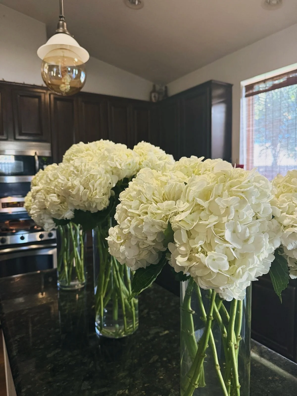 Three vases with white hydrangea flowers on a black marble kitchen countertop, dark wooden cabinets, microwave, light fixture, window with blinds in the background.