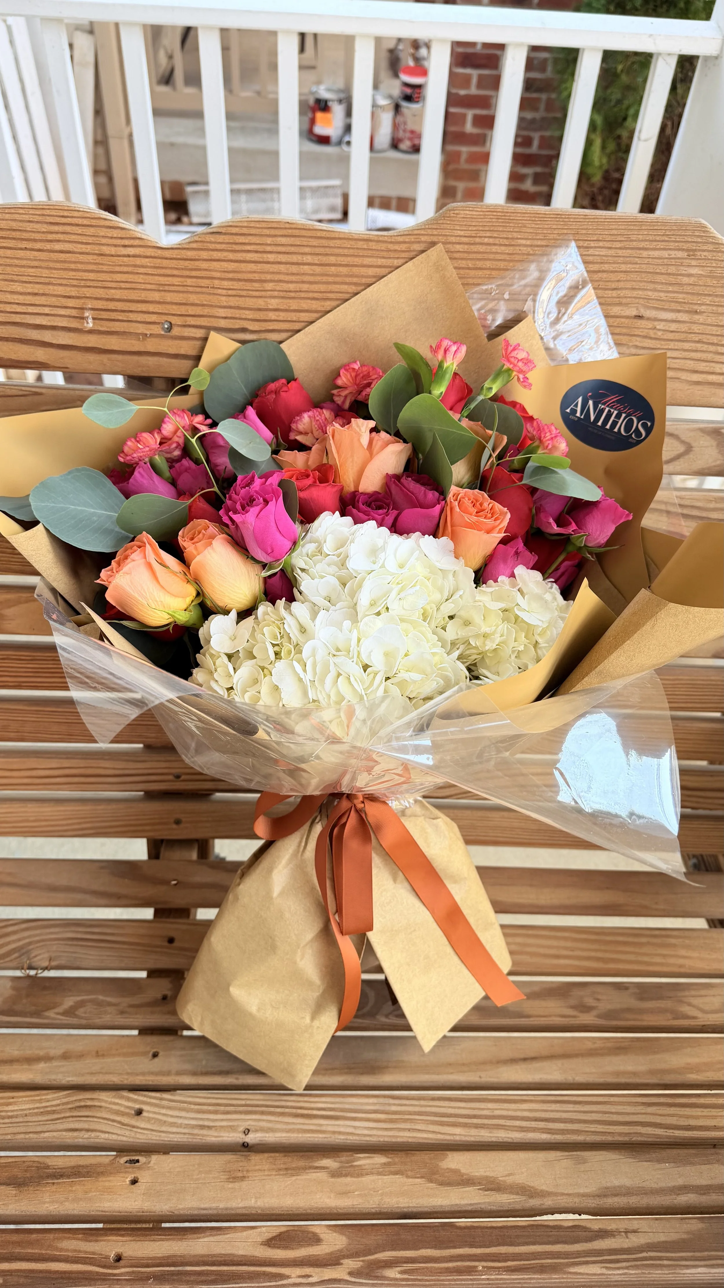 Colorful bouquet of mixed flowers including pink, orange, and white roses, pink carnations, and white hydrangeas, wrapped in brown paper and clear plastic, tied with an orange ribbon, placed on a wooden bench.