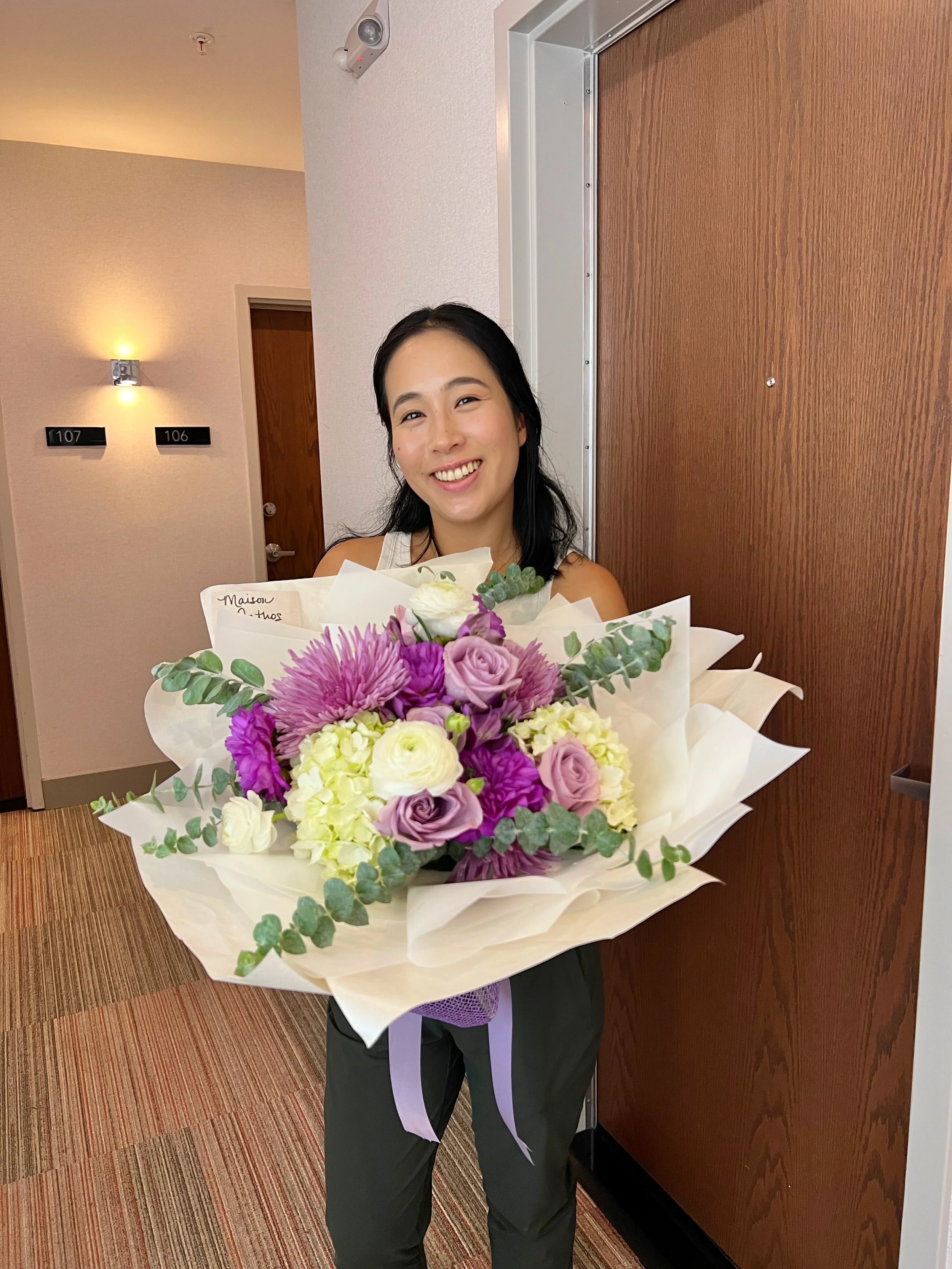 Young woman with black hair smiling and holding a large bouquet of colorful flowers, standing in a hallway near a wooden door.