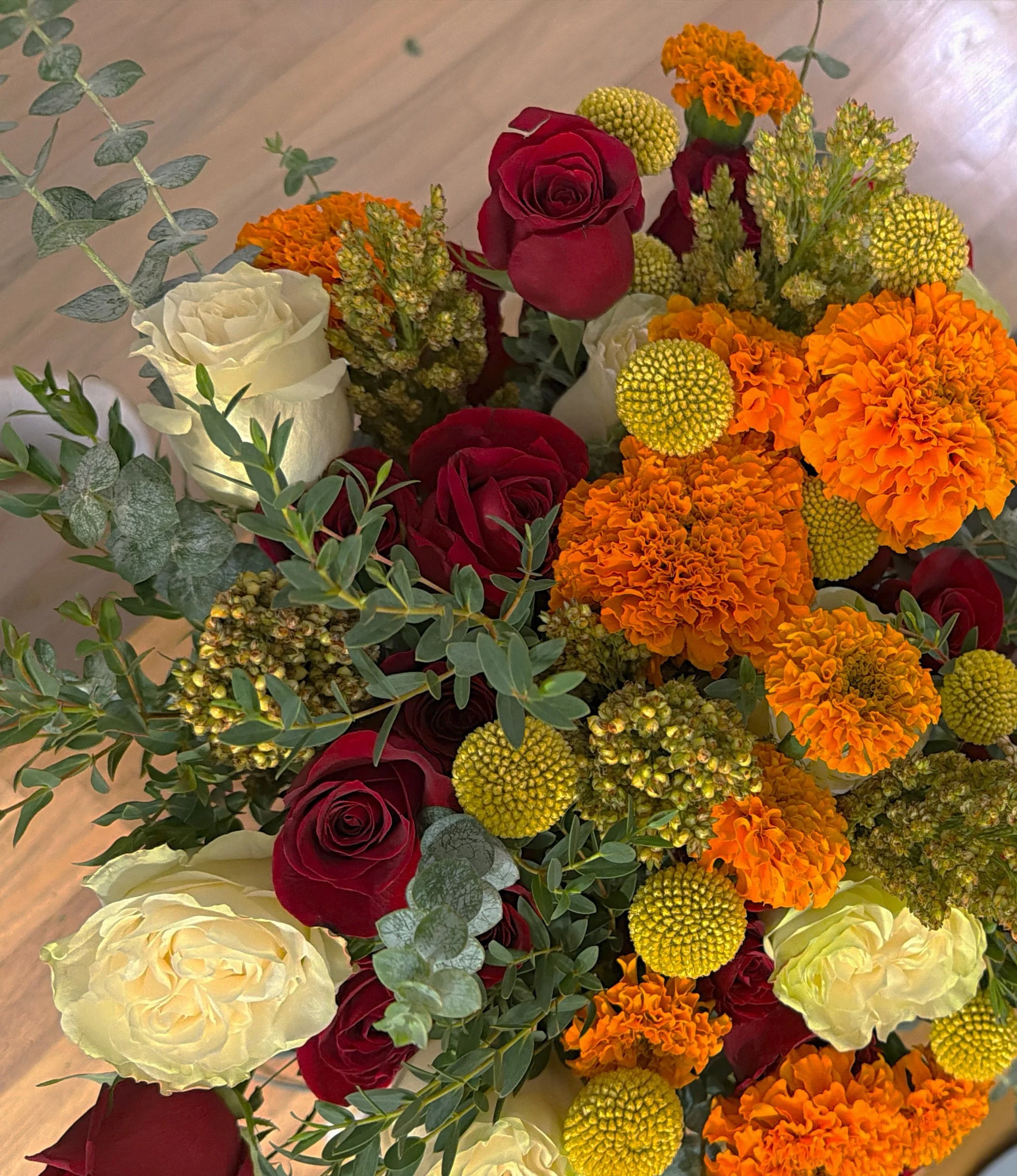 A bouquet of flowers featuring white roses, deep red roses, bright orange marigolds, and small yellow button flowers, accented with green leaves and eucalyptus.