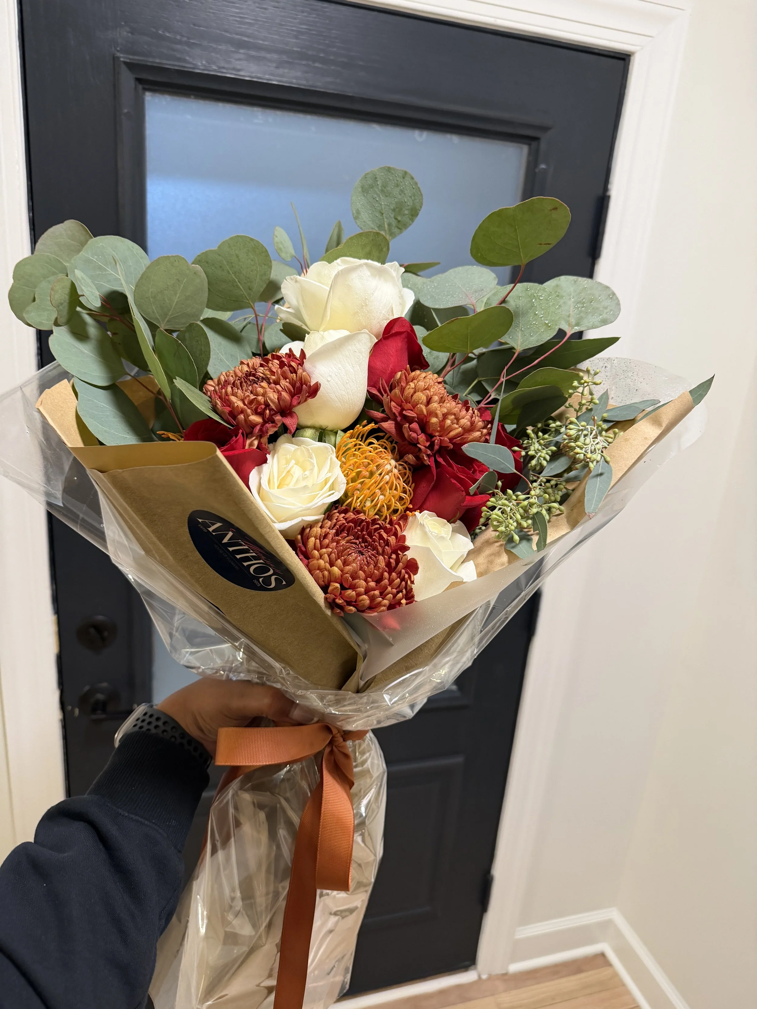 A hand holding a bouquet of flowers in front of a door. The bouquet includes white roses, red roses, orange and red chrysanthemums, a yellow pin cushion protea, and eucalyptus leaves, wrapped in brown paper and clear plastic with a brown ribbon.