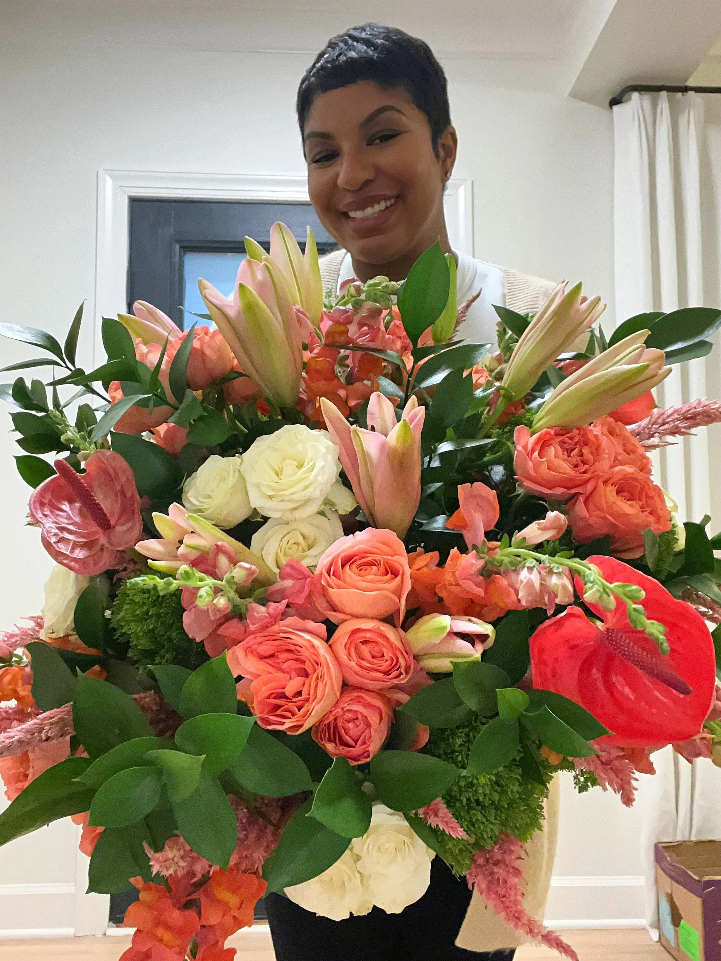 A woman holding a large, colorful bouquet of flowers, smiling, indoors with a white door, beige walls, and curtains in the background.