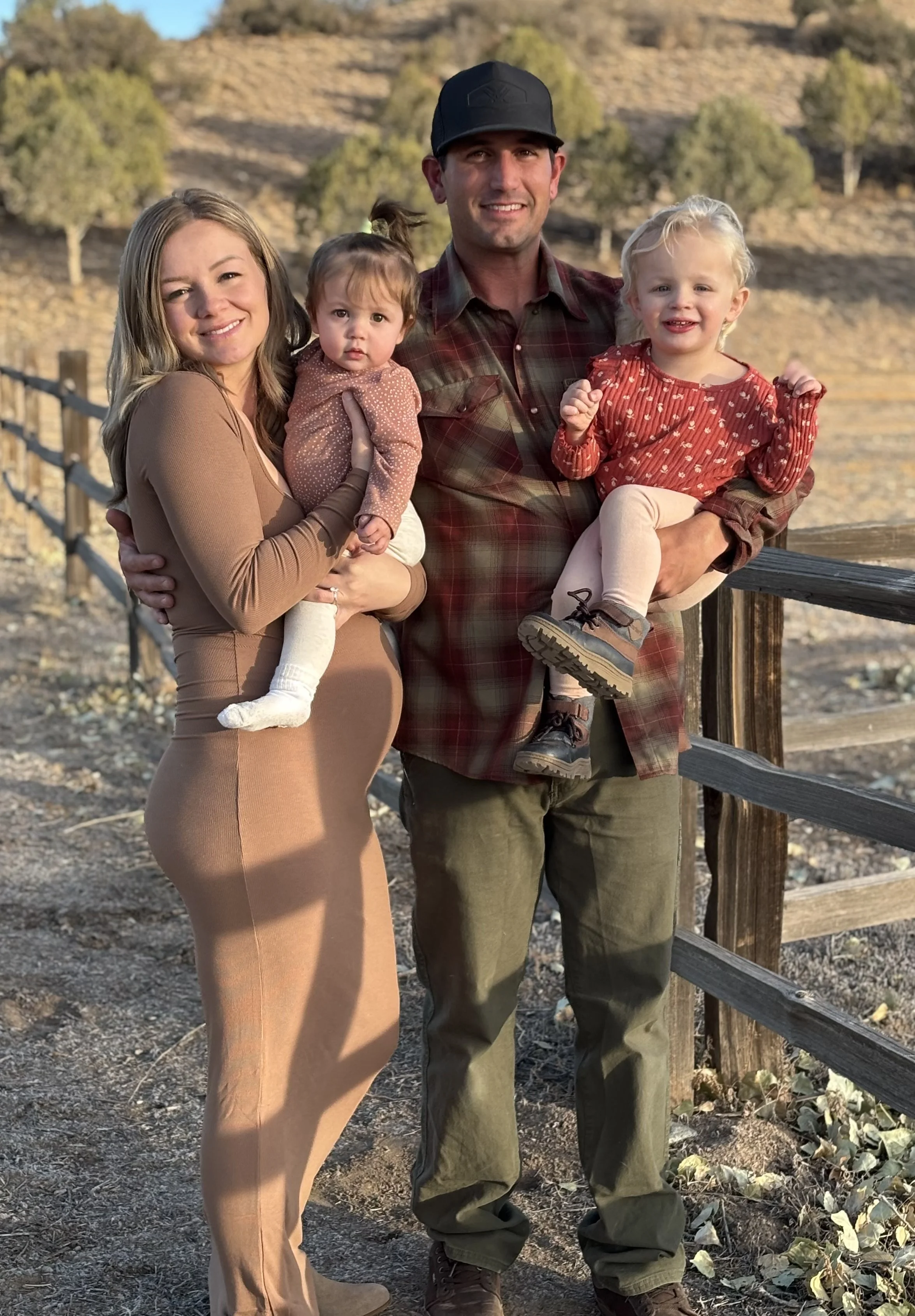 A family of four outdoors on a sunny day, with a wooden fence and hilly landscape in the background. The woman and man are smiling, each holding a young girl in their arms.