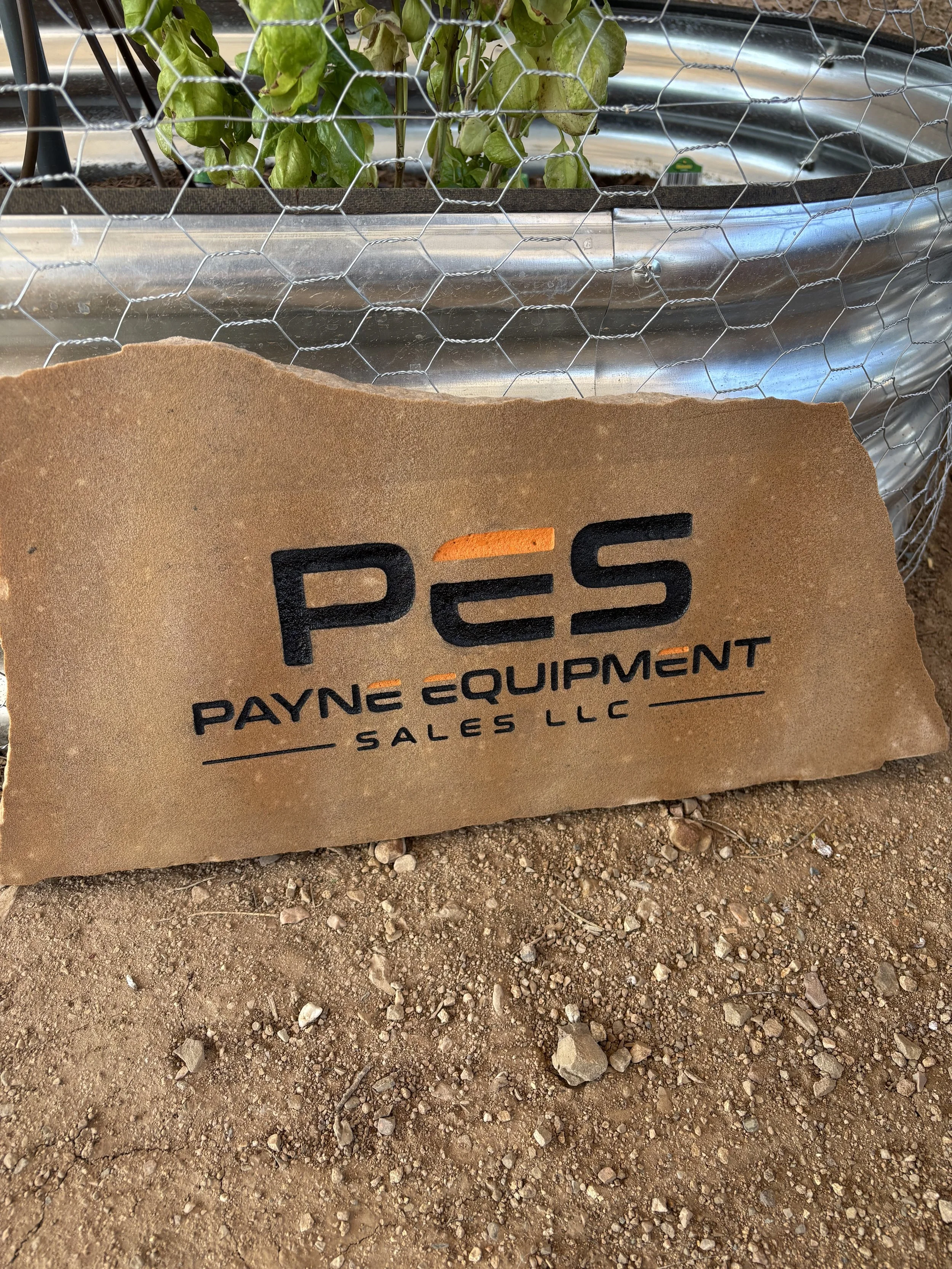 A tan stone sign with black and orange lettering reads "PES Payne Equipment Sales LLC". Behind it, there is a galvanized metal planter with green leafy plants, protected by a chicken wire fence, and the ground is dirt with small rocks.