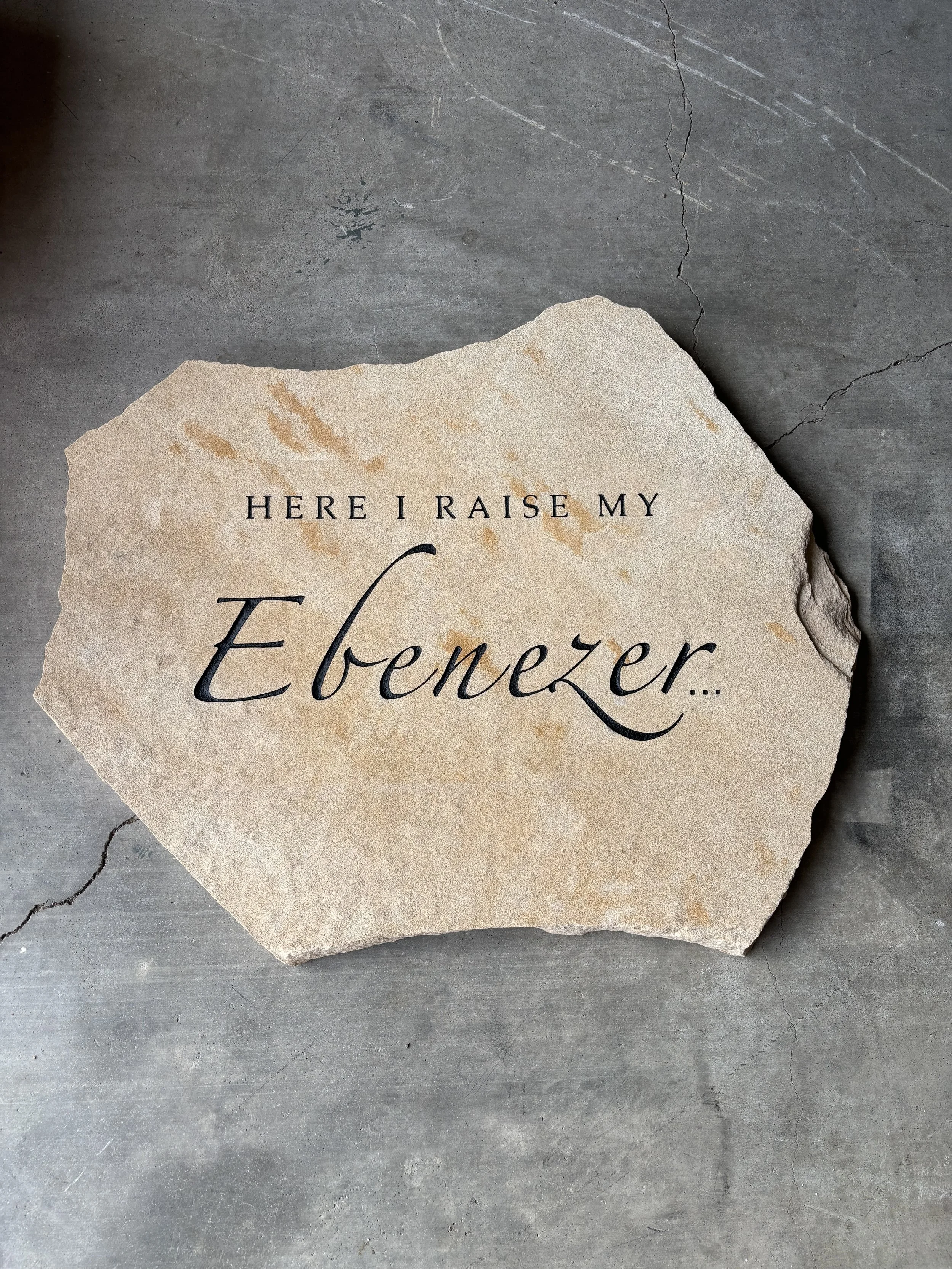 A piece of beige rock with the phrase 'Here I raise my Ebenezer...' written on it, placed on a gray concrete surface.