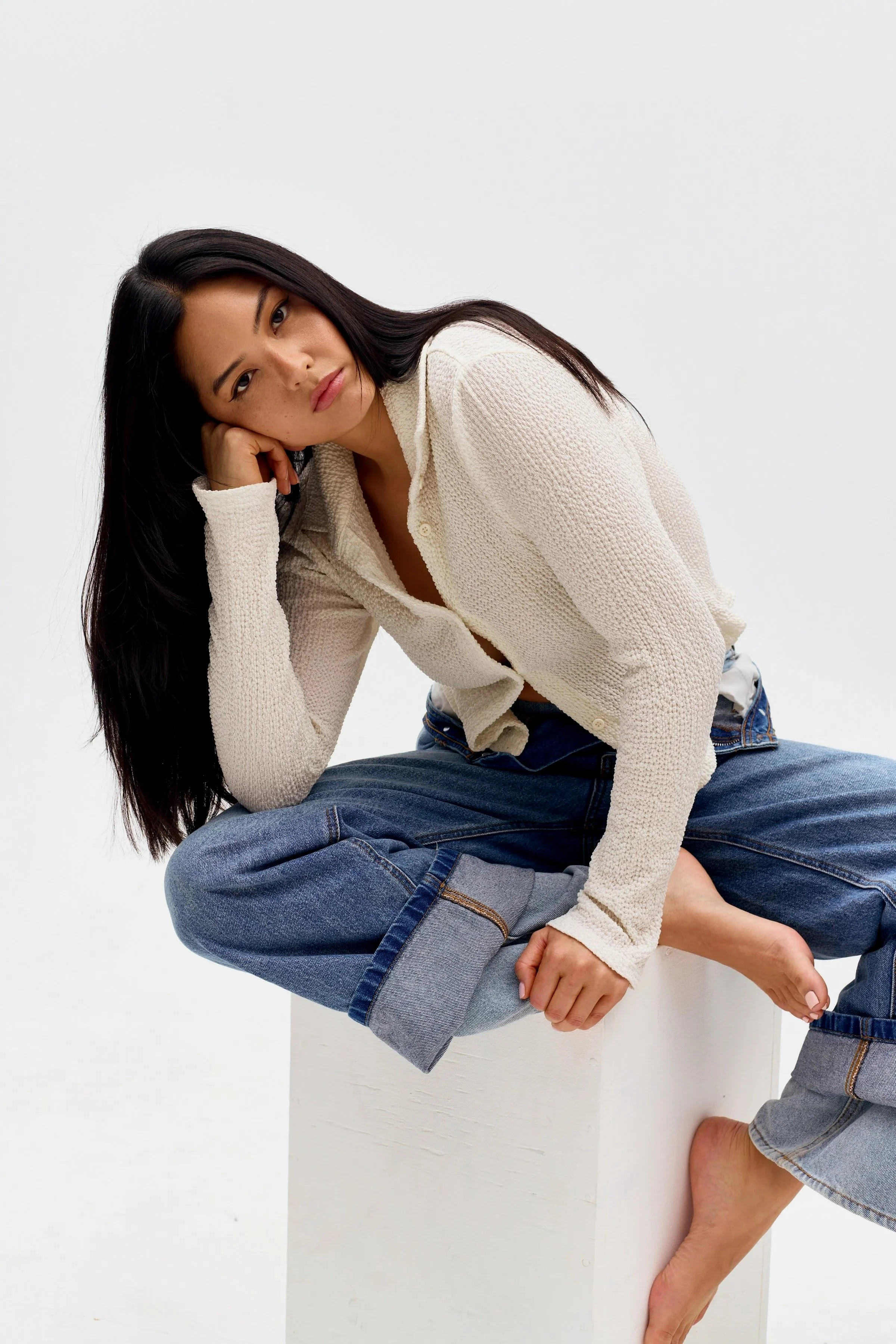 Woman with long dark hair sitting on a white block, wearing a cream-colored cardigan and rolled-up jeans, with a white background.