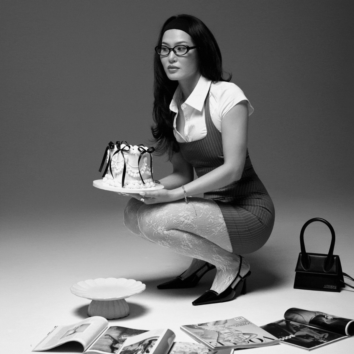 A woman with glasses wearing a white blouse, a striped skirt, and lace tights crouches holding a decorated cake. She is in a studio with magazines and a handbag on the floor.
