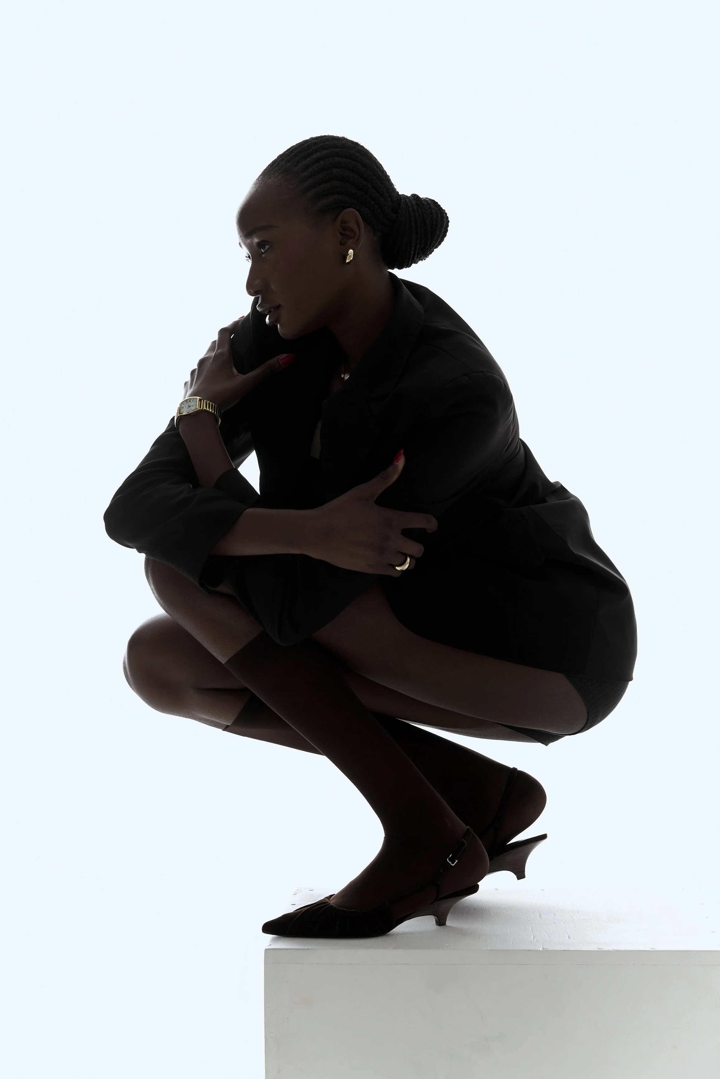 Woman squatting on a white block, dressed in dark clothing and high heels, holding a phone to her ear against a light background.