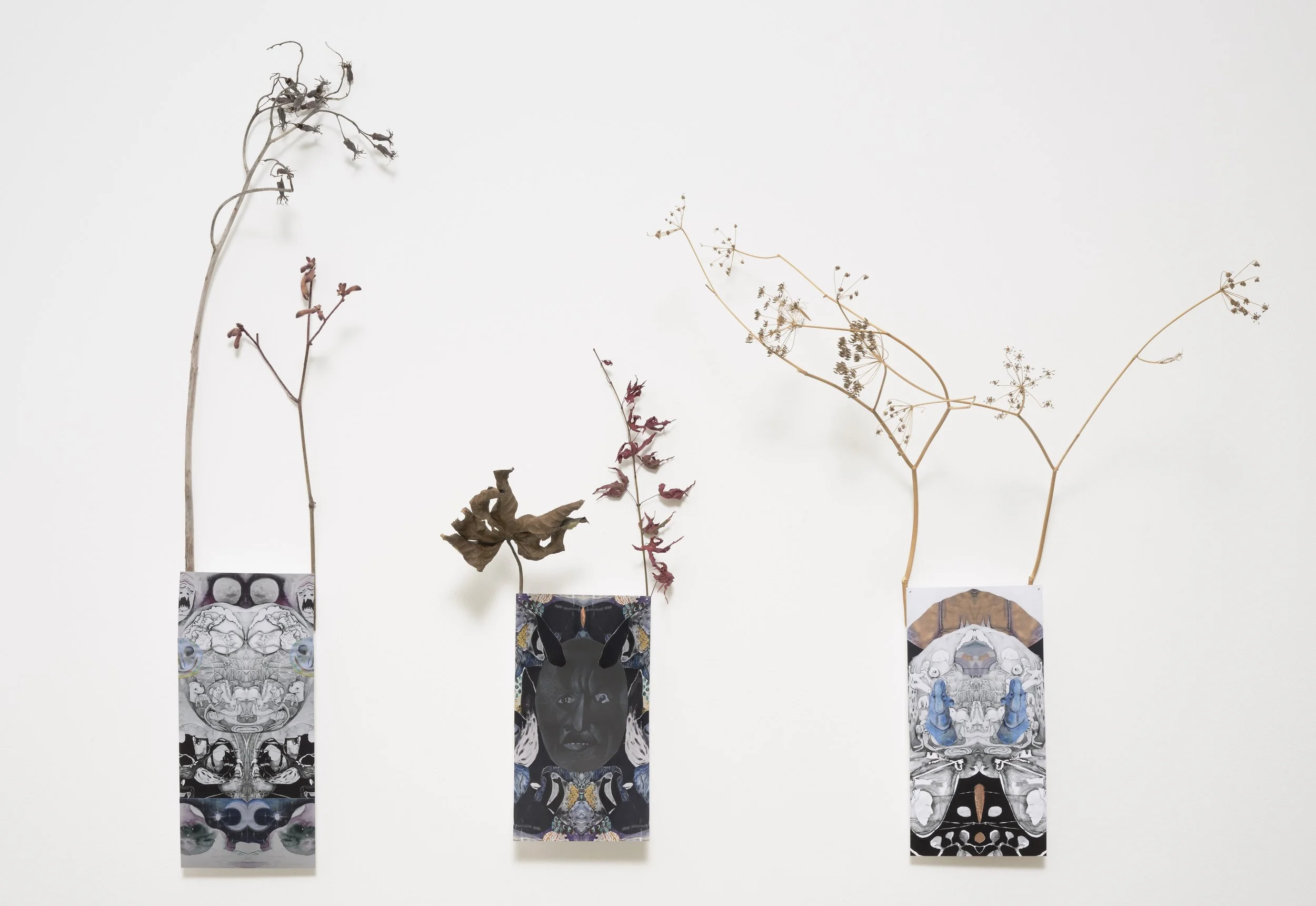 Three artworks with dried leaves attached on top, displayed on a white wall. Each art features a collage or drawing, with the leaves creating a three-dimensional extension.