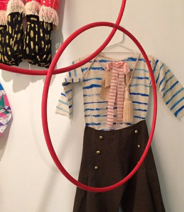 Display of a child's nautical-themed outfit, including a striped shirt, brown skirt with gold buttons, pink striped scarf, and beige tassel, hung on a white wall with a red hula hoop around it.