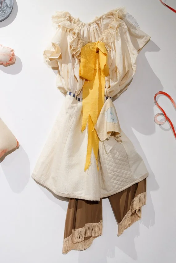 A vintage-style dress with a yellow ribbon at the waist, beige puffed sleeves, and tan fringe-trimmed pants attached at the bottom, displayed on a white wall.