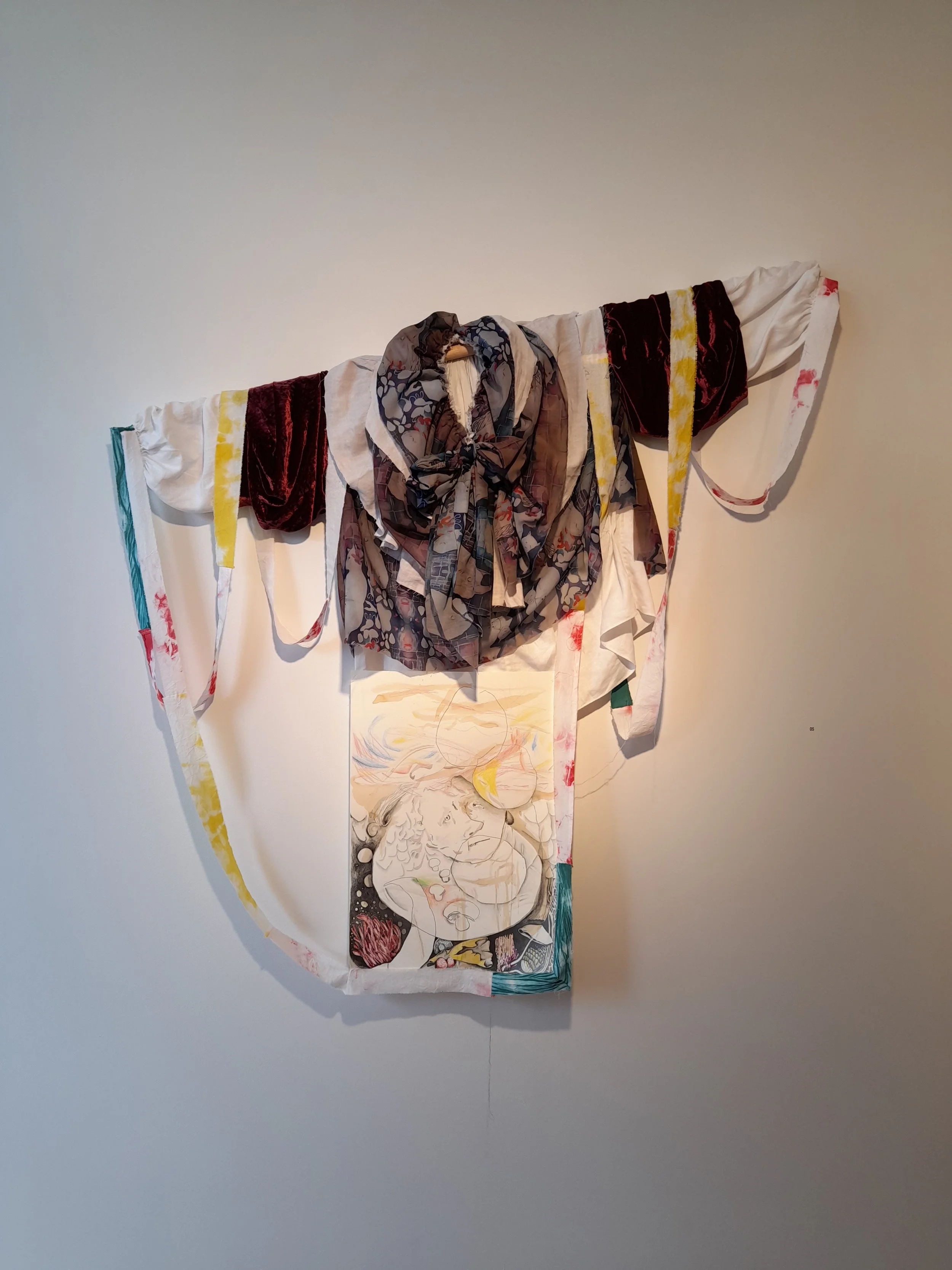 Mixed media artwork on a white wall, featuring a fabric with a large scarf draped over the top, colorful abstract lines, and a sketch of a woman's face.
