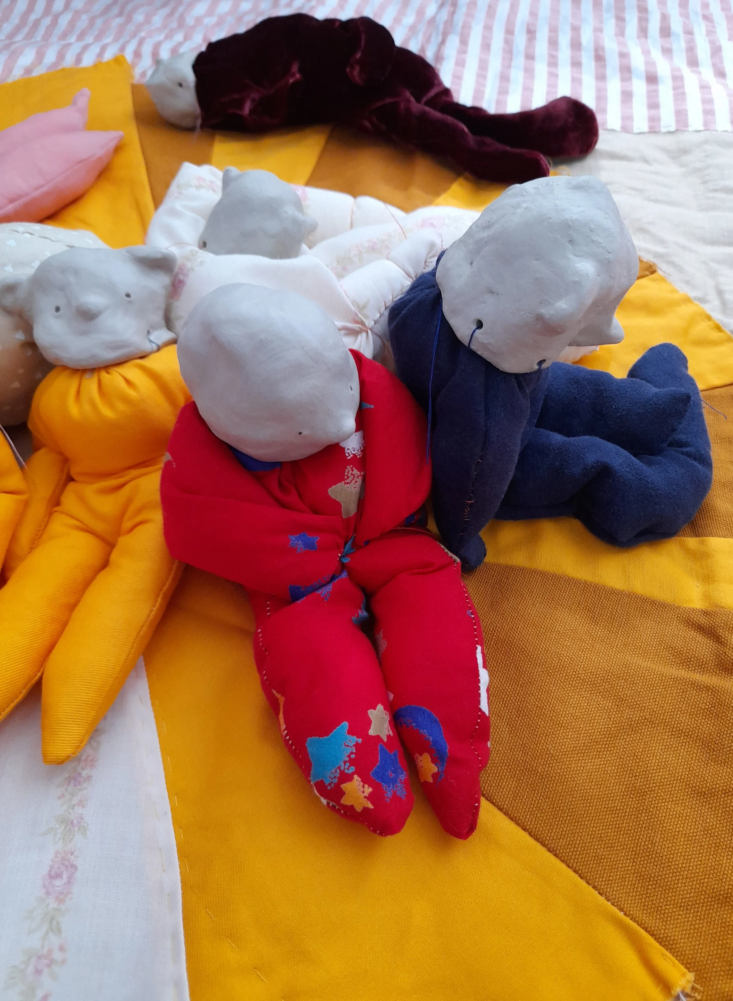 Collection of handmade fabric and paper mache puppets resembling animals and people, arranged on a bed with yellow and pink bedding.