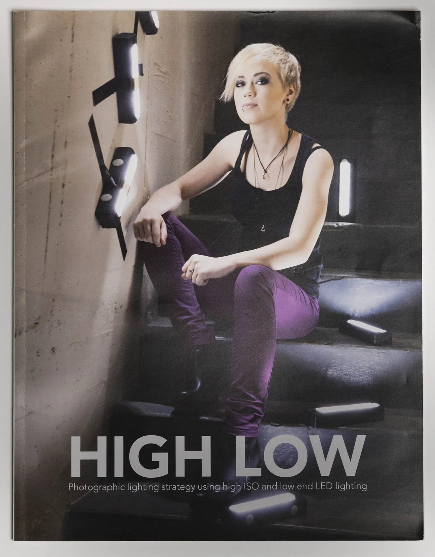 Photo of a young woman with short blonde hair, wearing a black tank top and purple pants, sitting on dark stairs with LED lights on the wall next to her. The text on the photo reads 'HIGH LOW' and a subtitle discusses photographic lighting strategy.