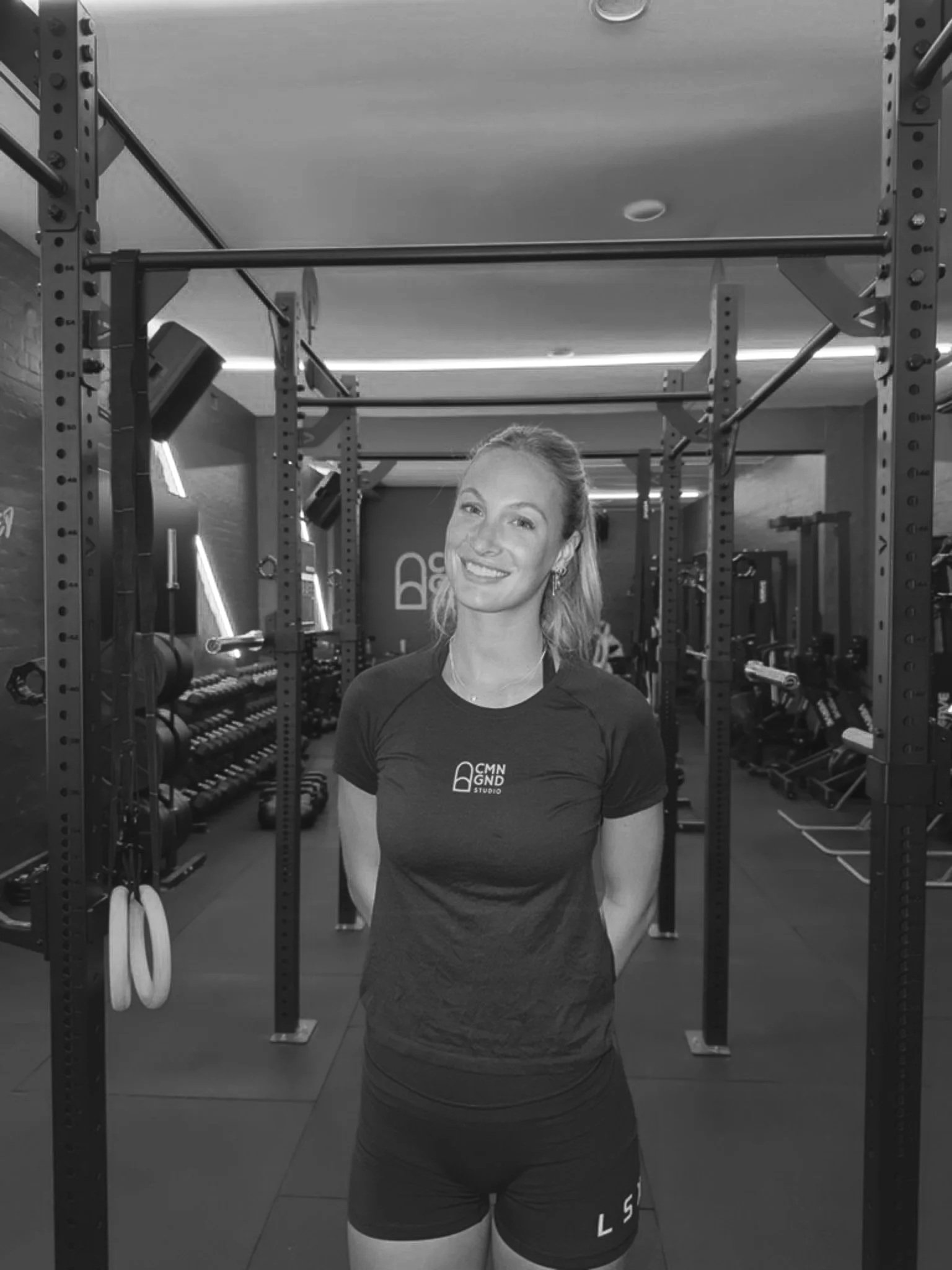A woman smiling at the camera in a gym, standing behind a power rack with workout equipment in the background.