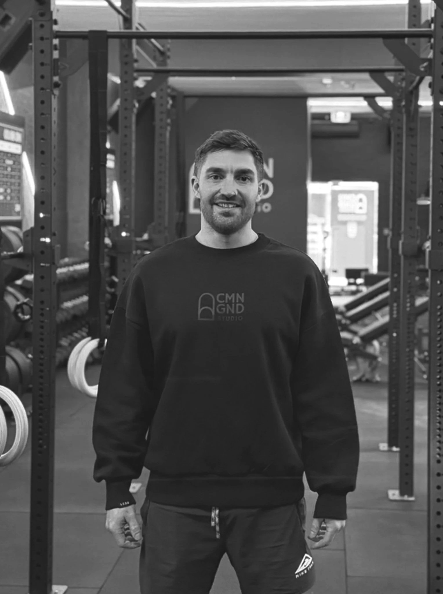 A man standing in a gym with workout equipment and weights in the background, smiling and wearing a sweatshirt with 'CMN GND Studio' logo.