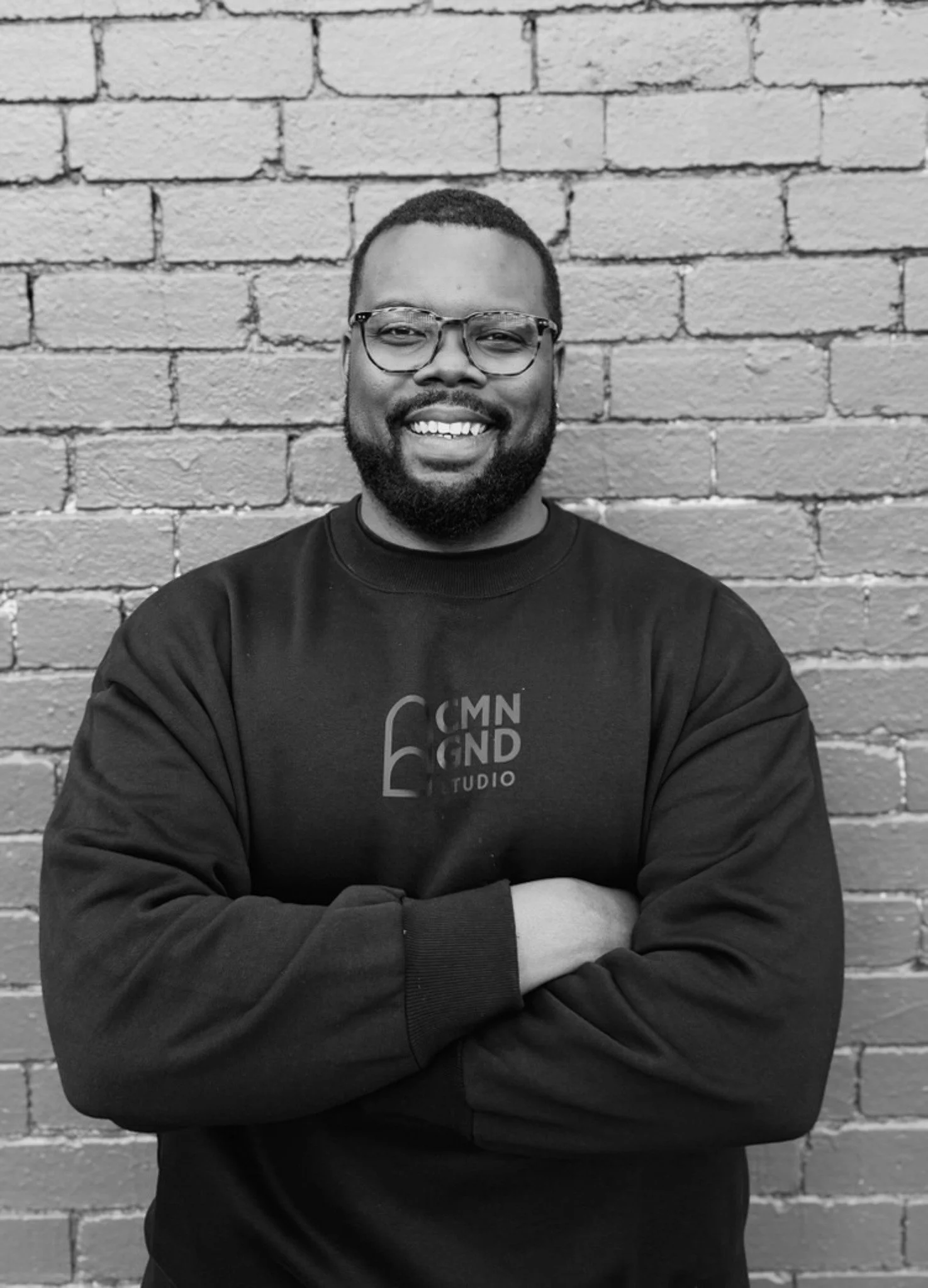 A smiling man with glasses and a beard, wearing a black sweatshirt with the logo '6 CMN GND studio', standing against a brick wall with arms crossed.