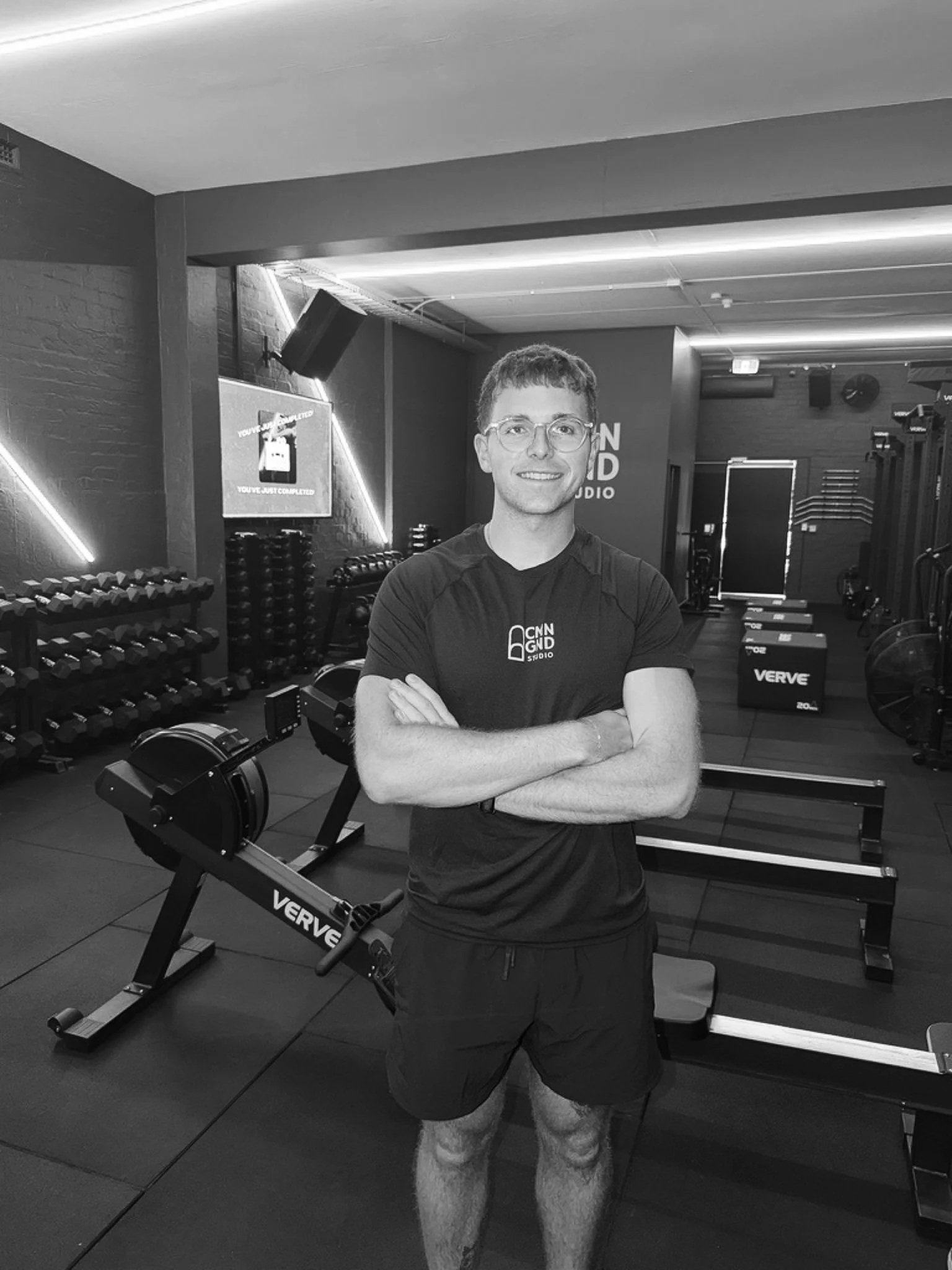 A young man with glasses and short hair standing with arms crossed in a gym, surrounded by workout equipment including dumbbells, rowing machines, and plyometric boxes.