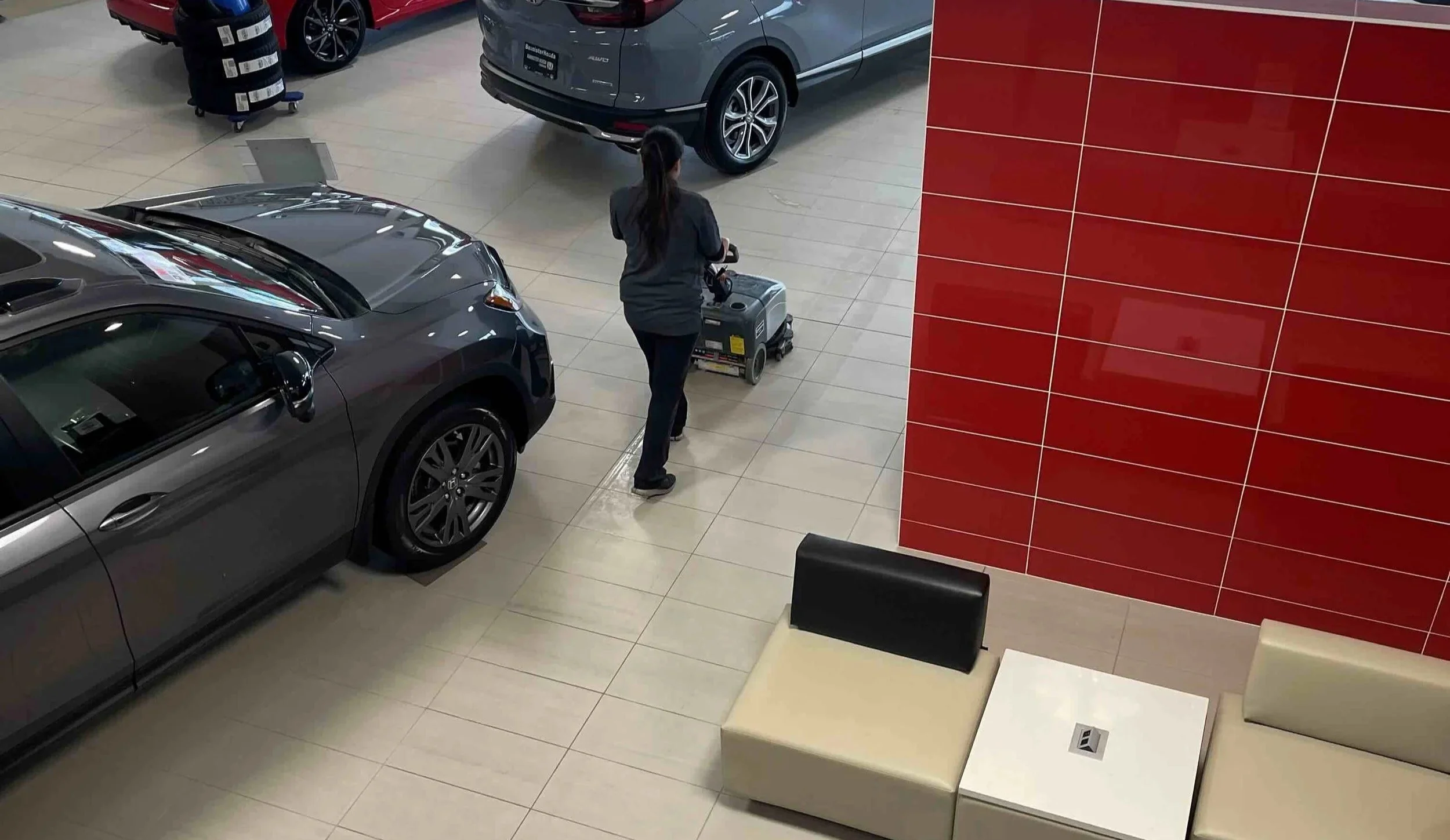 Buzz Brite Cleaning Solutions team member using a floor cleaning machine in a car dealership showroom with several cars and a red tiled wall.