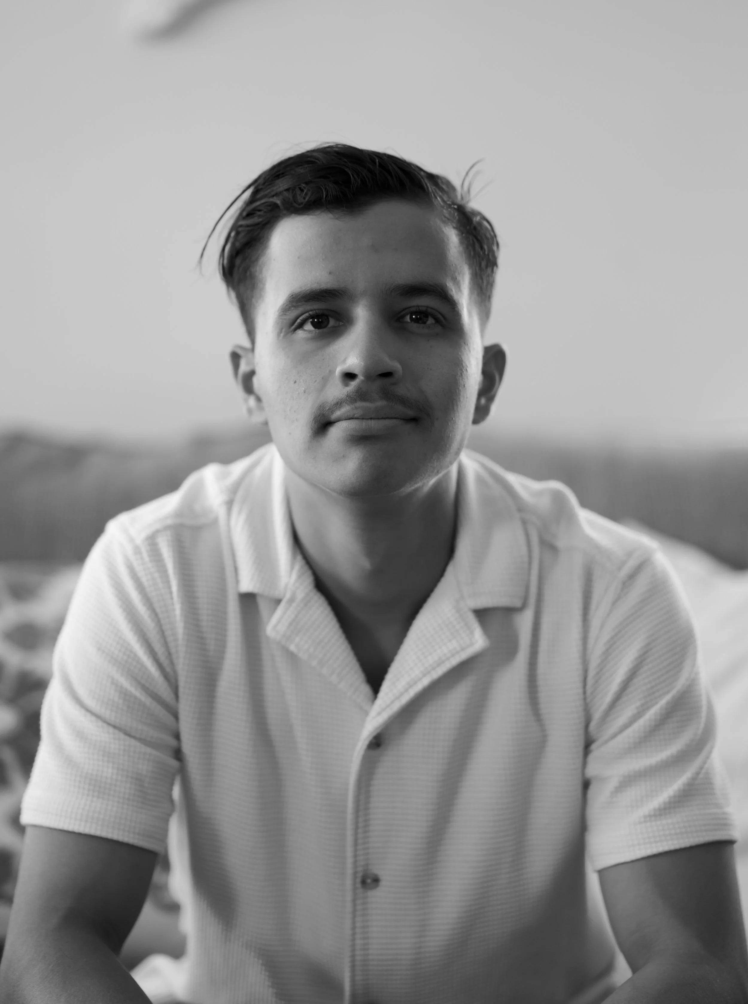A young man with dark hair and a slight smile, wearing a short-sleeved, collared shirt, sitting indoors with a blurred background.