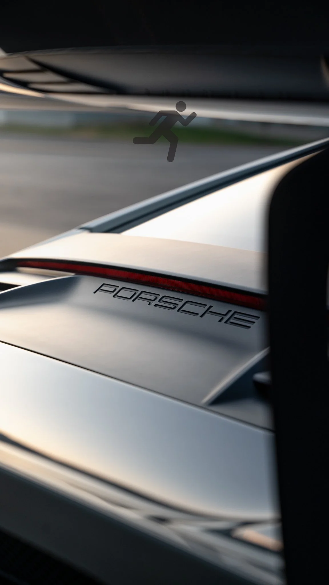 Close-up of a sports car with Porsche logo, part of the rear and spoiler visible, on a road during sunset.