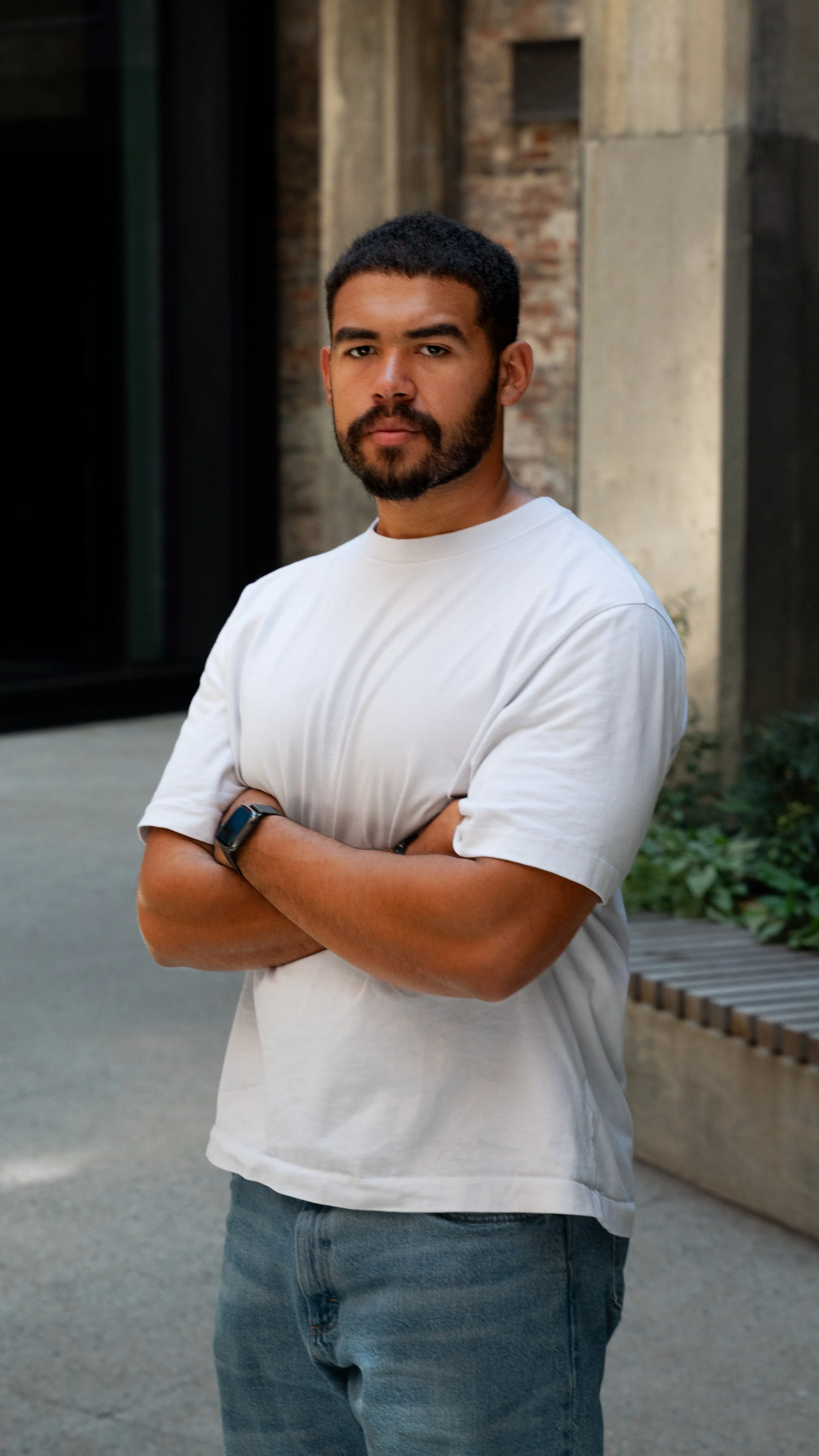 A young man with dark hair and a beard standing outdoors with arms crossed, wearing a white t-shirt, jeans, and a smartwatch on his left wrist, in front of a brick and concrete building.