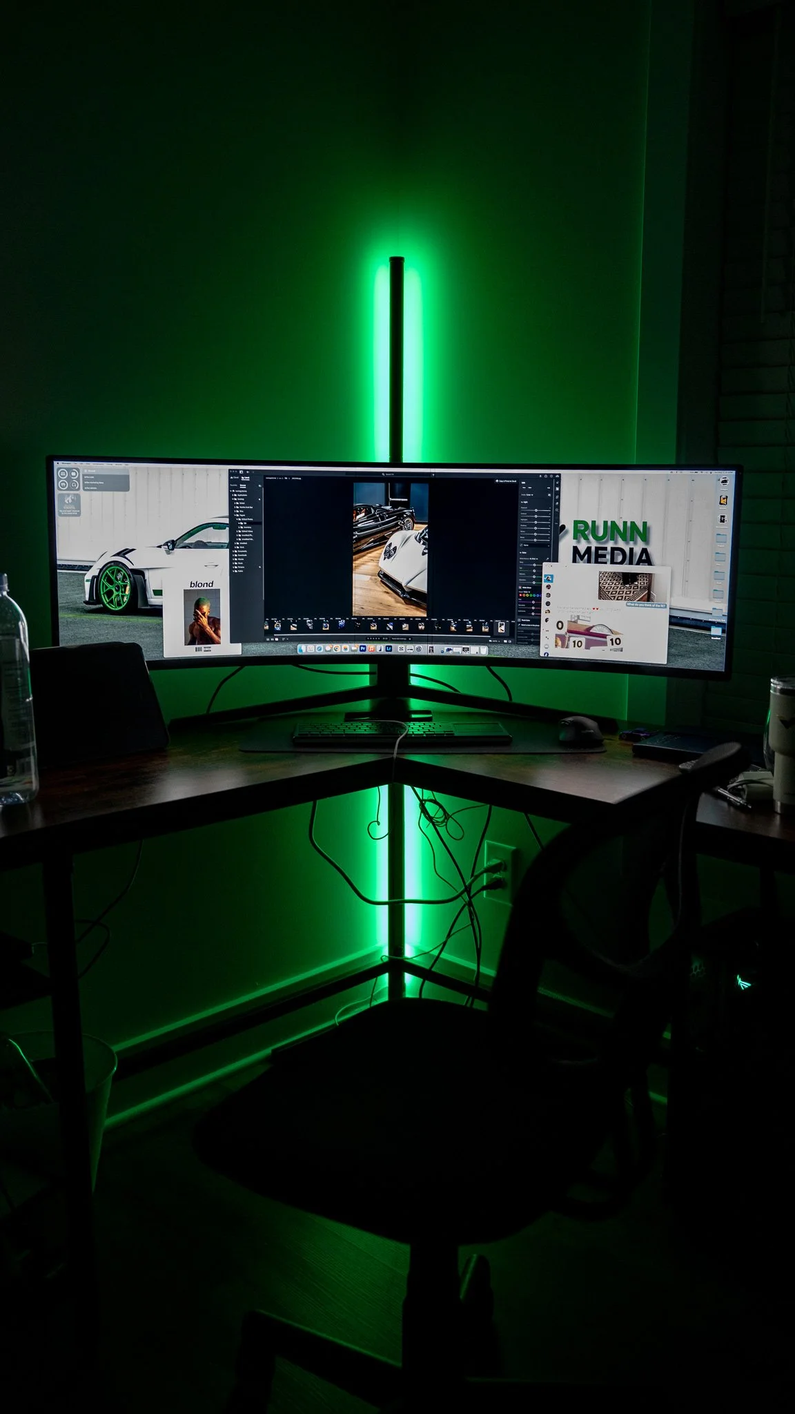 A computer desk with a large curved monitor displaying a photo editing app, with neon green LED light strip behind the monitor in a dark room.