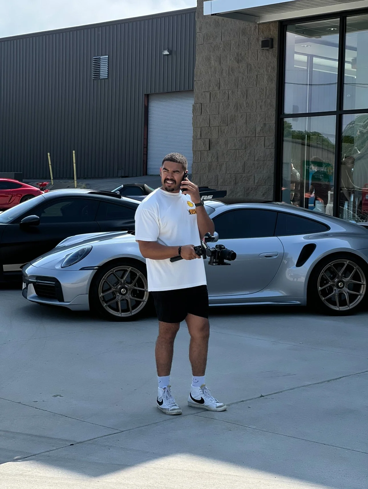 A man standing outdoors near a silver sports car, holding a camera in one hand and a phone to his ear in the other, with a black car behind him and a building with large windows nearby.