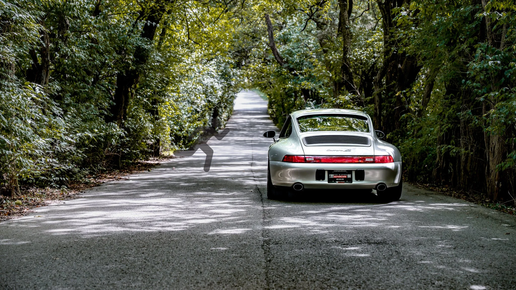 A silver Porsche 911 parked on a narrow tree-lined road with dappled sunlight and shadows.