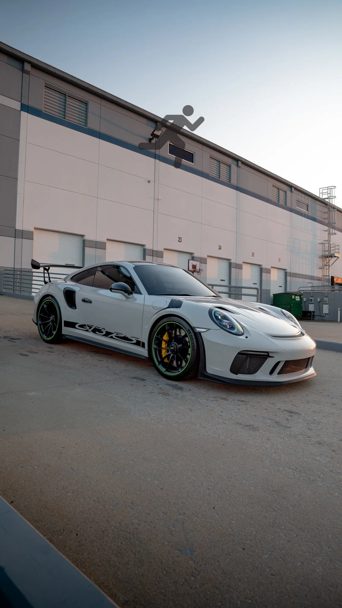 A white Porsche 911 GT3 RS sports car with black and green accents parked outside a warehouse during sunset.