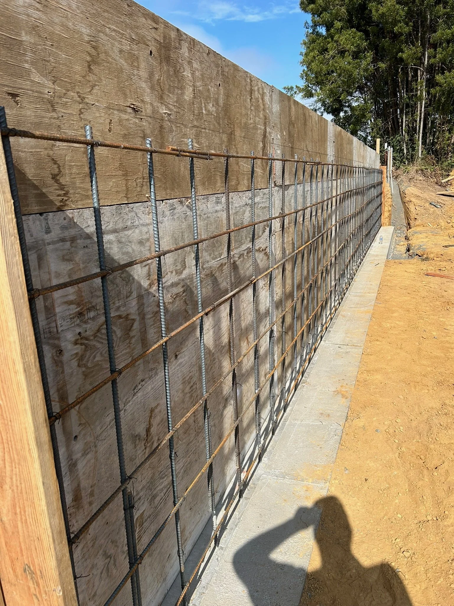 Construction site with rebar and wooden formwork along a concrete foundation, on a dirt area, with trees in the background and a blue sky.