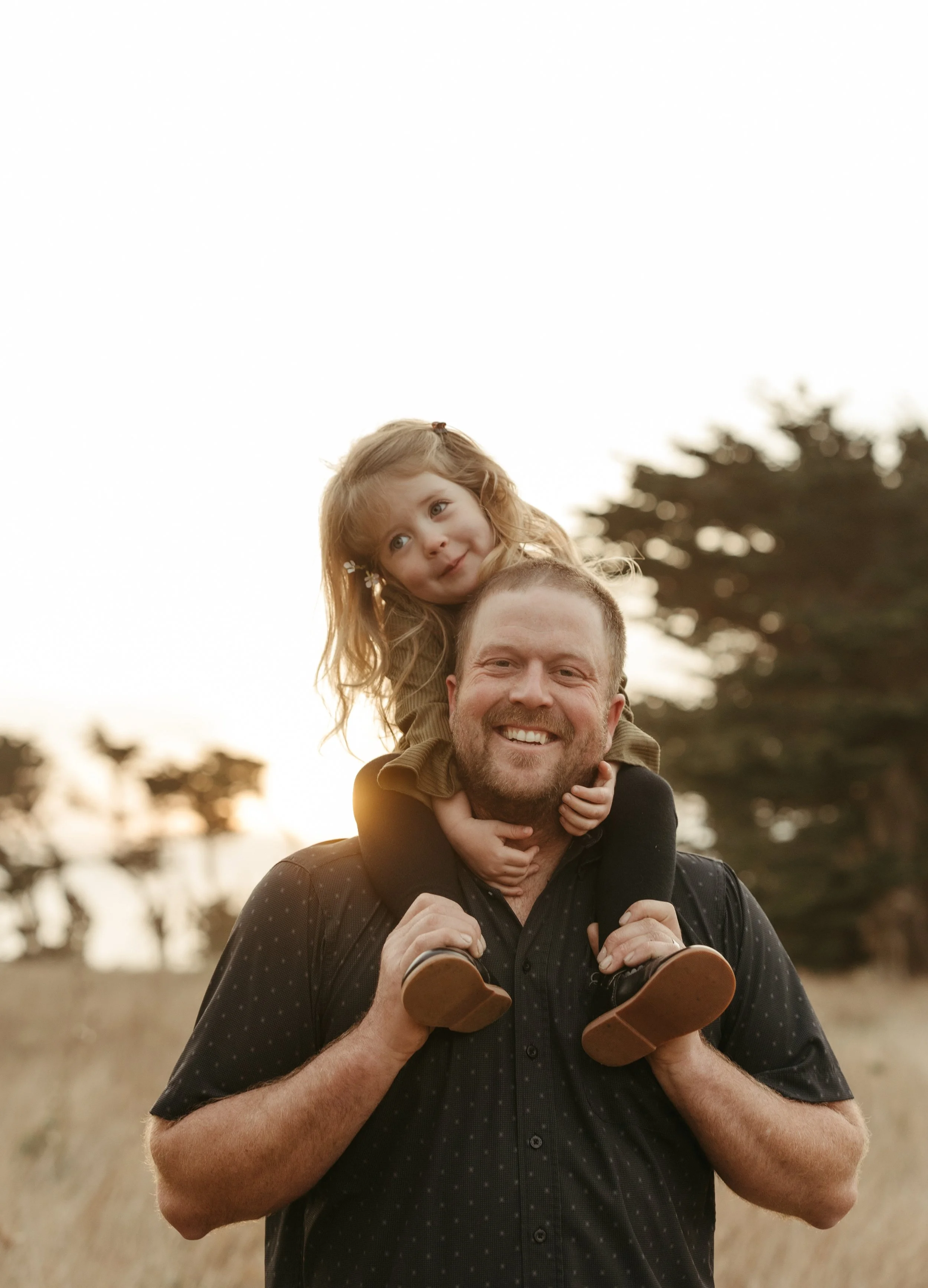 A man carrying a young girl on his shoulders outdoors during sunset, with trees in the background.