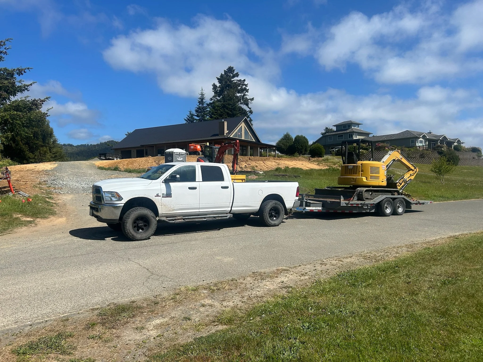A white pickup truck towing a trailer with a yellow excavator on it, parked on a suburban street with houses and trees in the background.