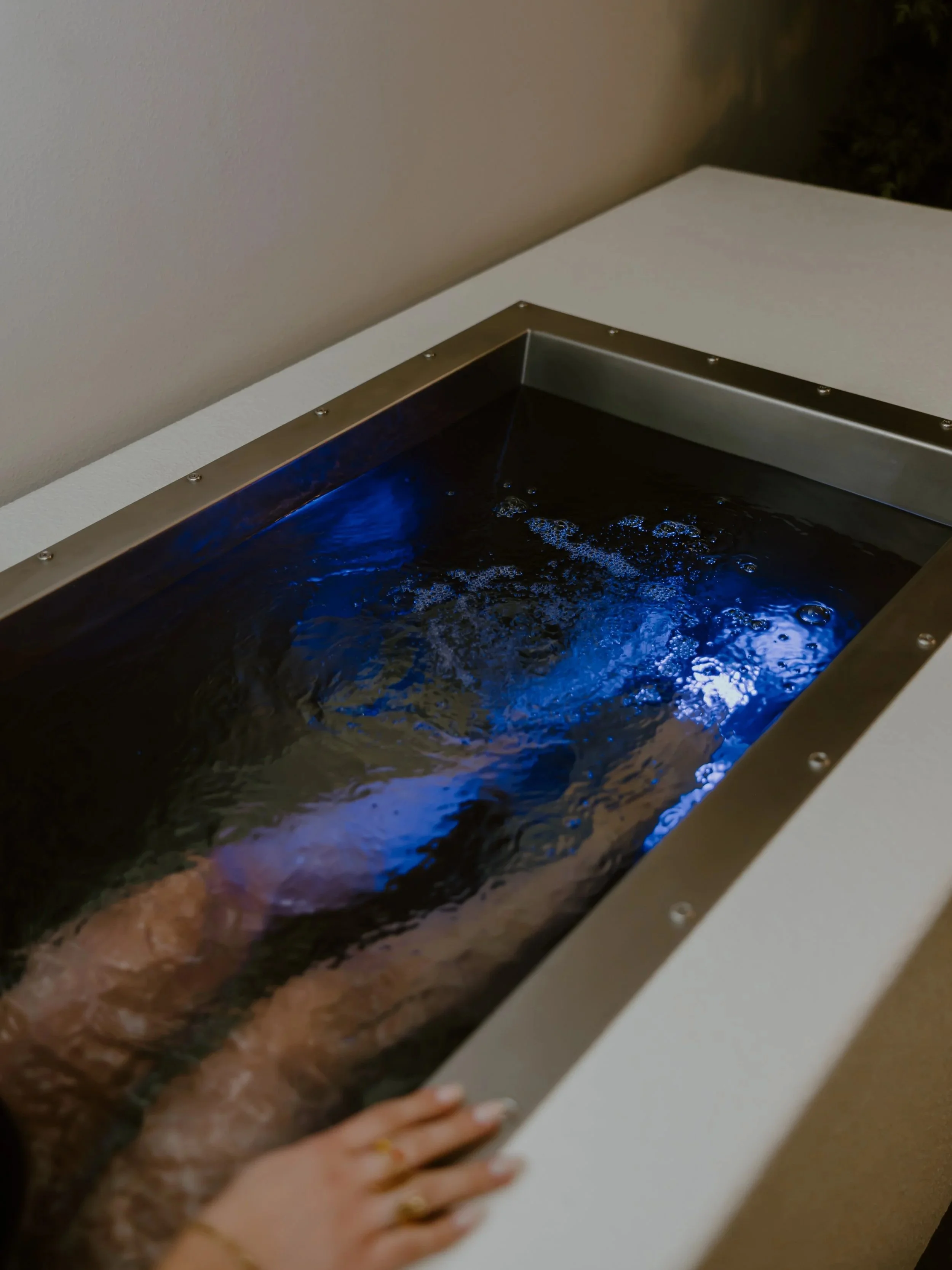 Person relaxing in a luxurious black soaking tub with blue underwater lights, located indoors with a beige wall and a plant visible nearby.