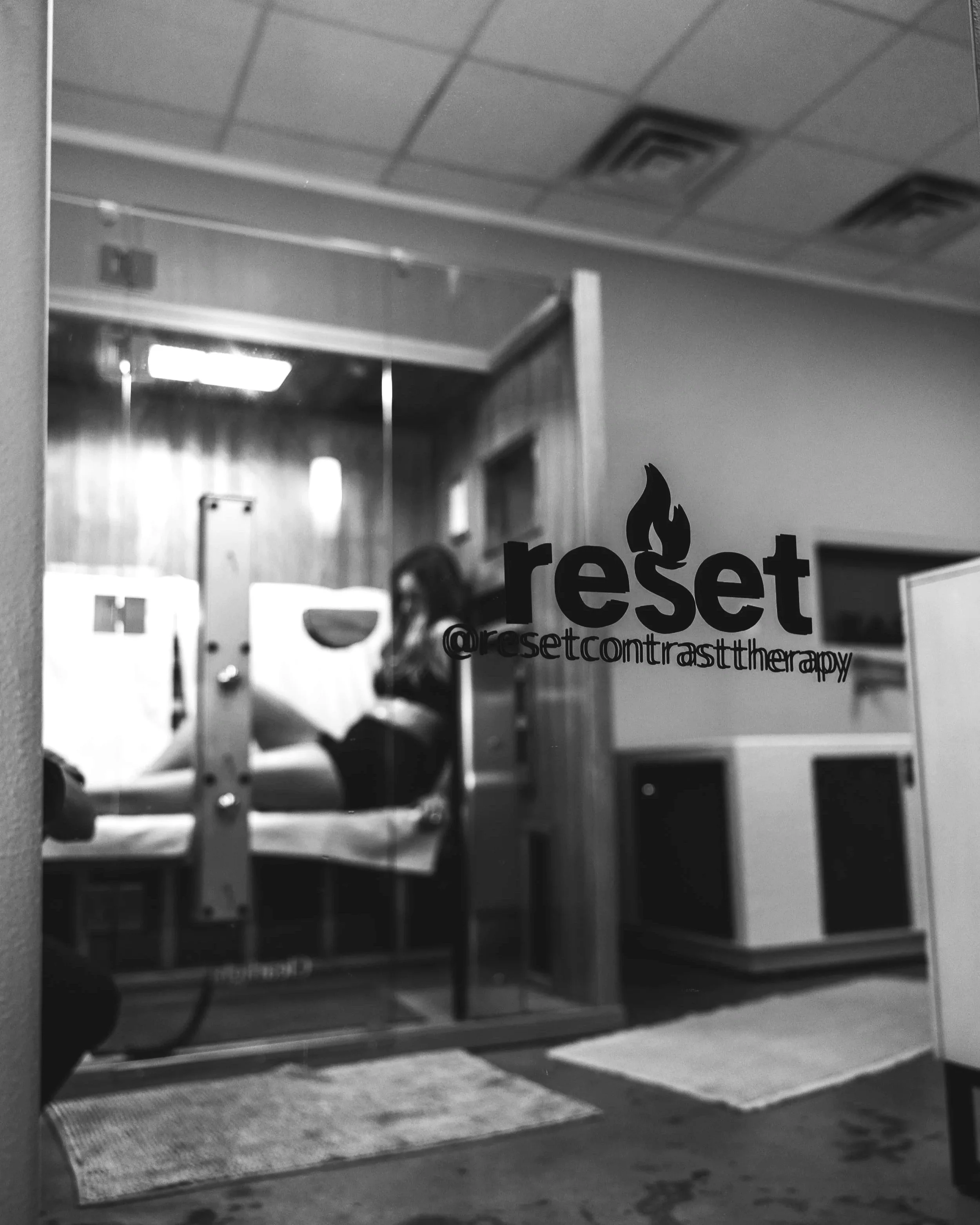 A woman sitting on a therapy bed through a glass door at a therapy center named 'Reset'.