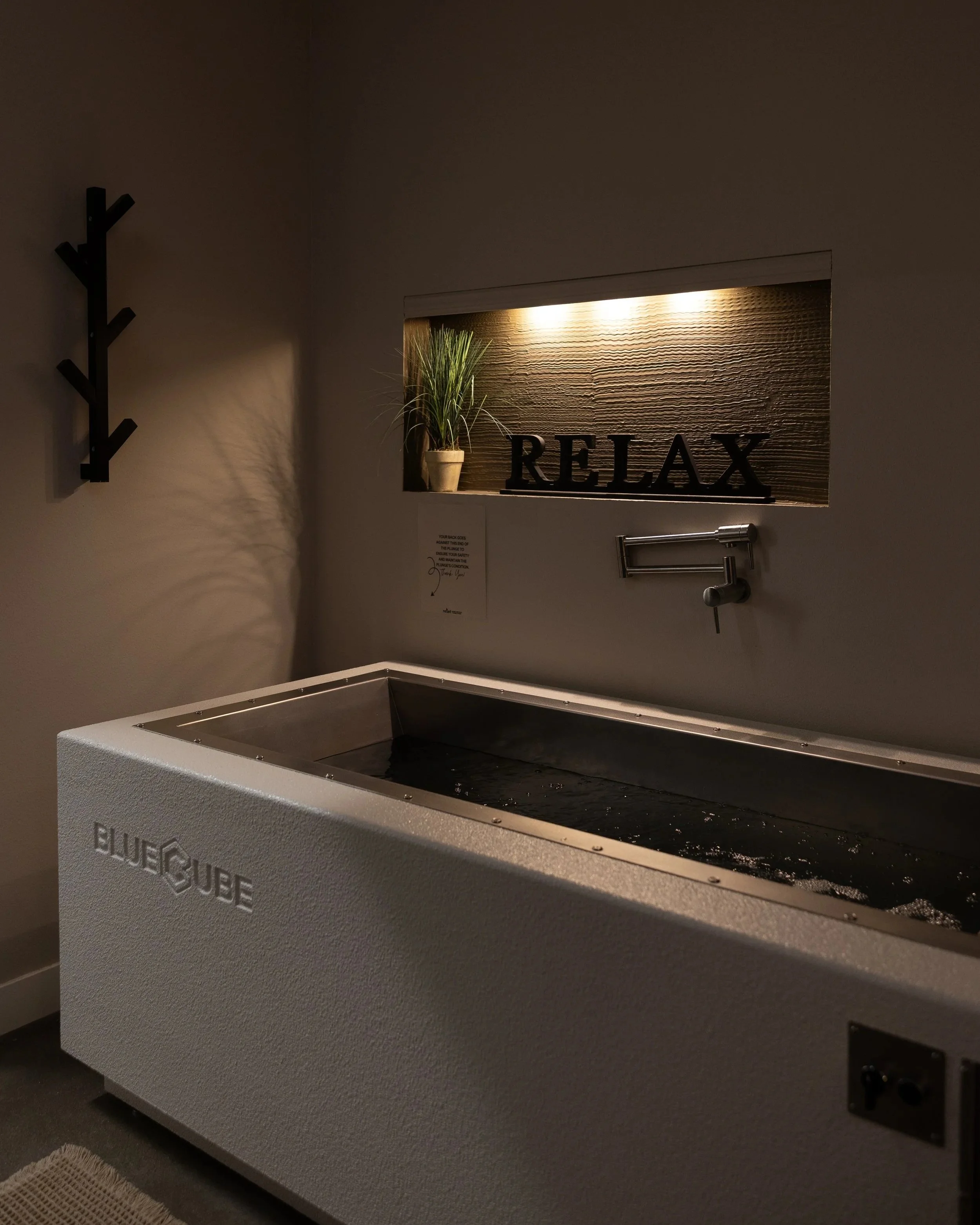 A white Bluecube hot tub in a room with minimalist decor, a wall niche with a potted plant, lighting, and a sign that says 'RELAX'.