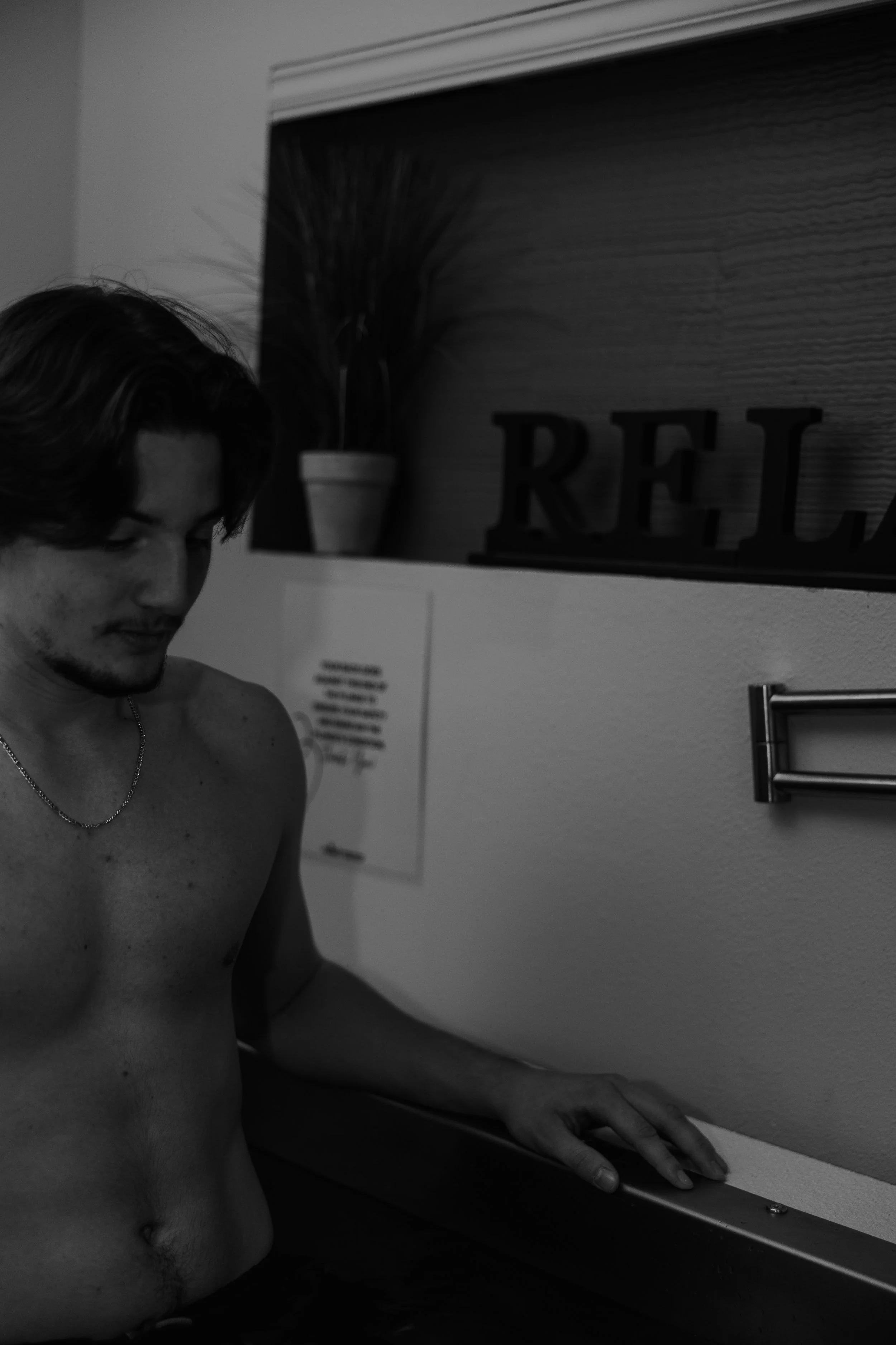 A shirtless young man with shoulder-length hair holding onto a surface indoors, with a potted plant and a sign that reads 'REL' in the background.
