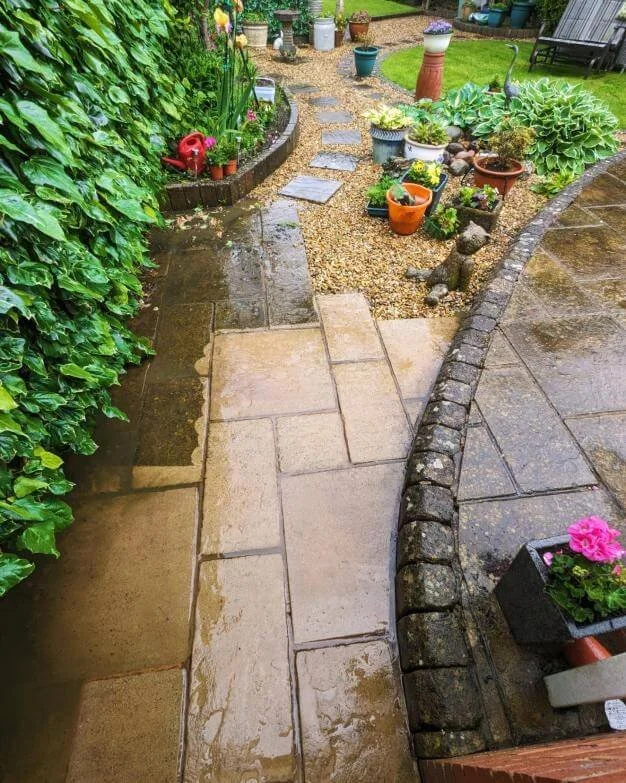 Wet garden pathway with stepping stones, potted plants, and lush green foliage on the left side, with a gravel and brick border on the right side, partially cleaned.