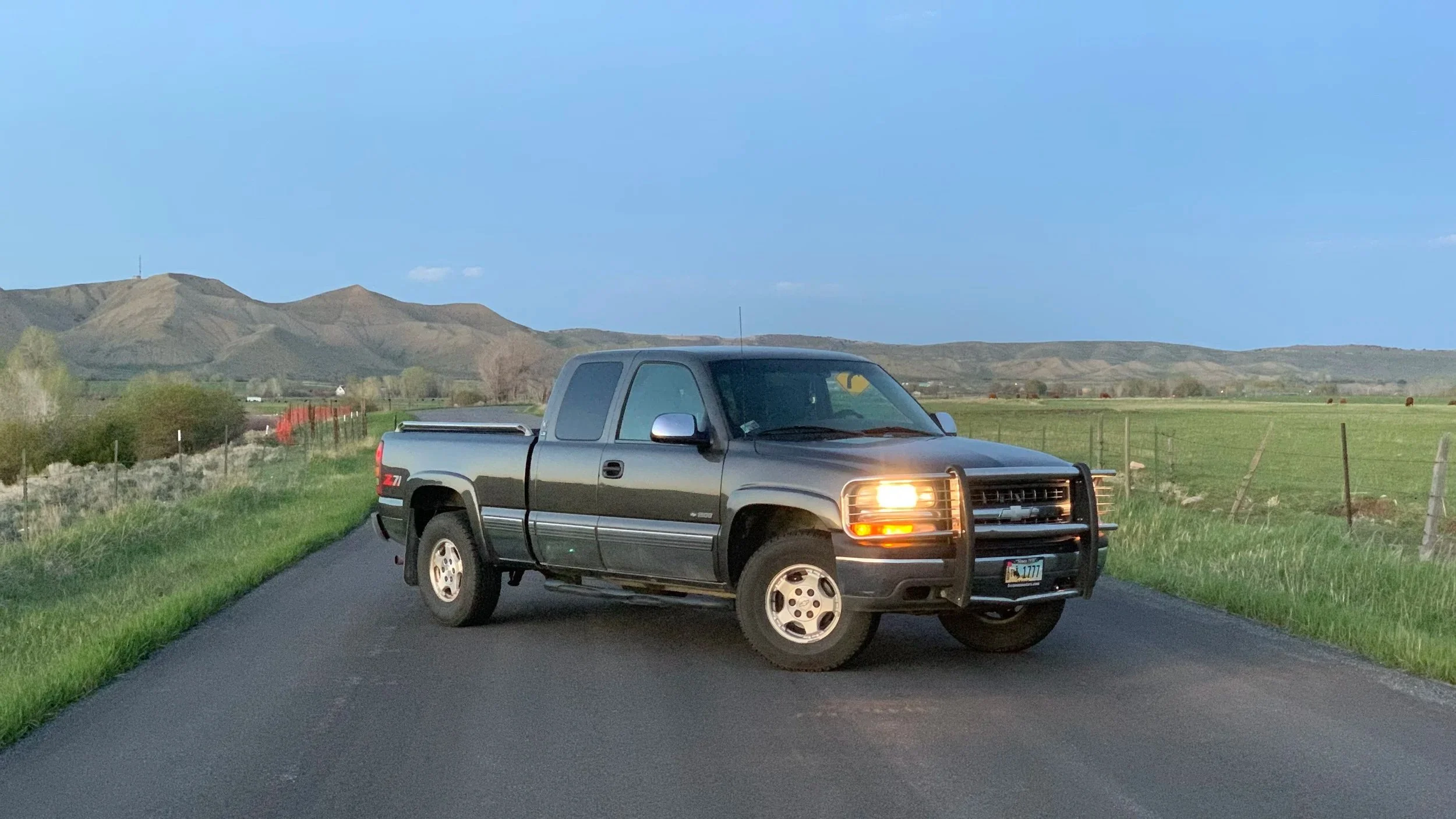 A black Chevrolet pickup truck with its headlights on parked diagonally on a rural paved road, surrounded by green grass, with open fields and rolling hills in the background under a clear blue sky.