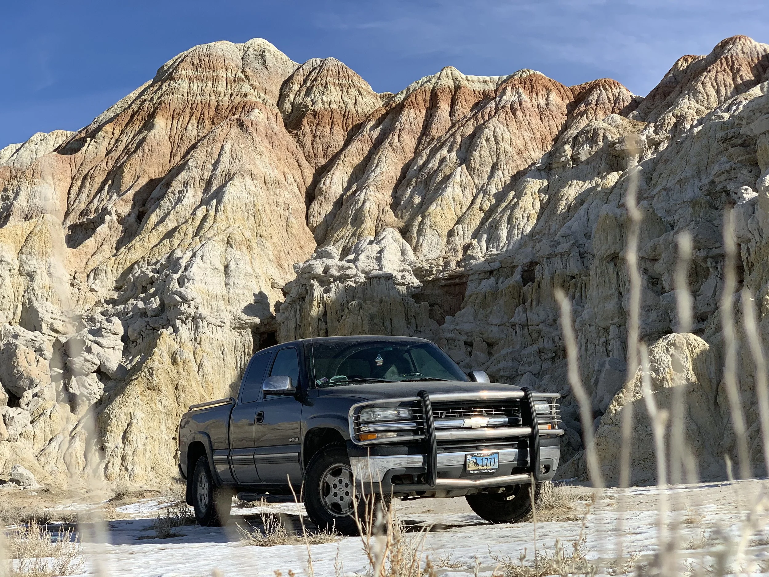 A silver pickup truck parked on a snowy landscape with rocky, multicolored mountains and a clear blue sky in the background.