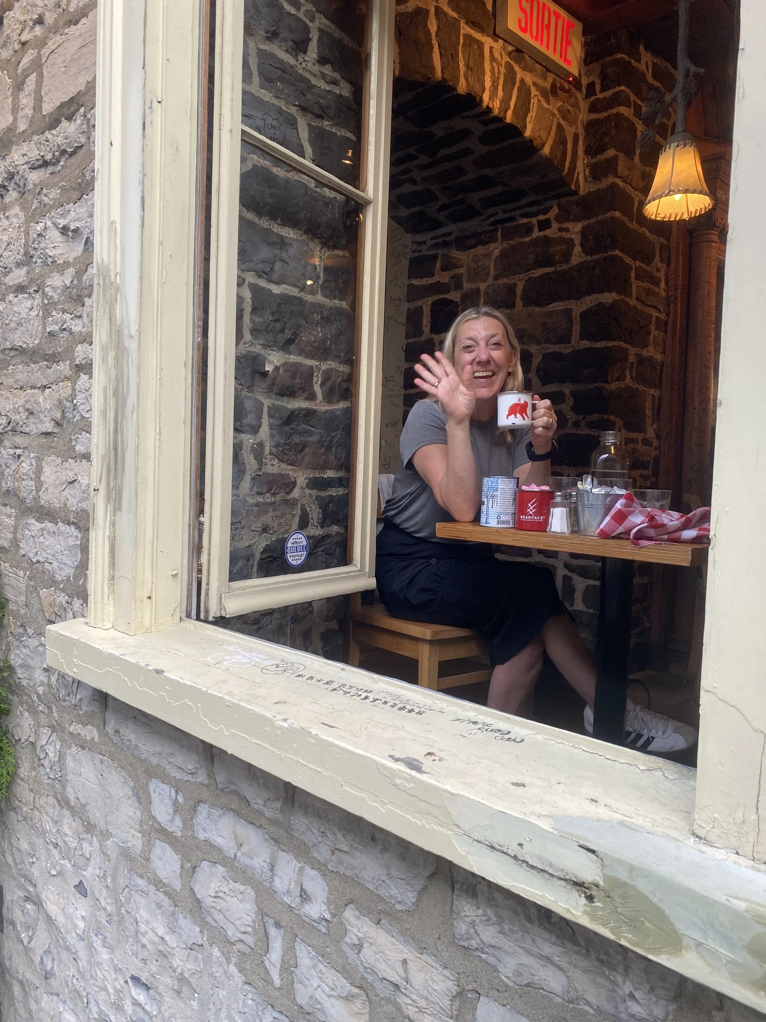 A woman sitting at a small table in a stone-walled cafe, smiling and waving while holding a white mug with a red pig logo. There are some items on the table, including a glass jar, a cup, and a checkered cloth. The window frame and interior lighting give the scene a cozy atmosphere.