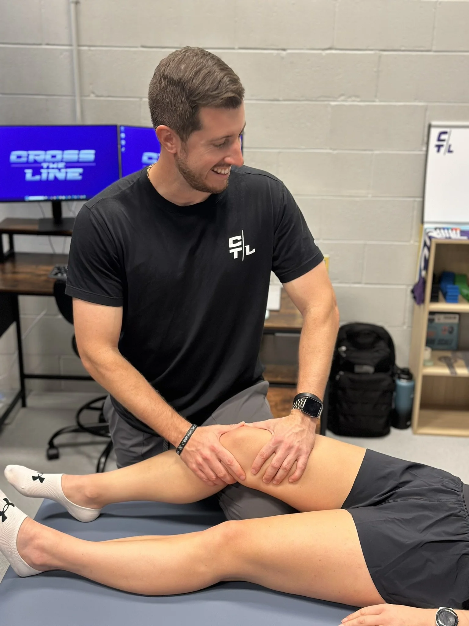 A man performs manual therapy interventions on a person's knee as part of a sports physical therapy session while smiling. The person receiving treatment is lying on a table, wearing gray shorts and white socks, with their legs extended.