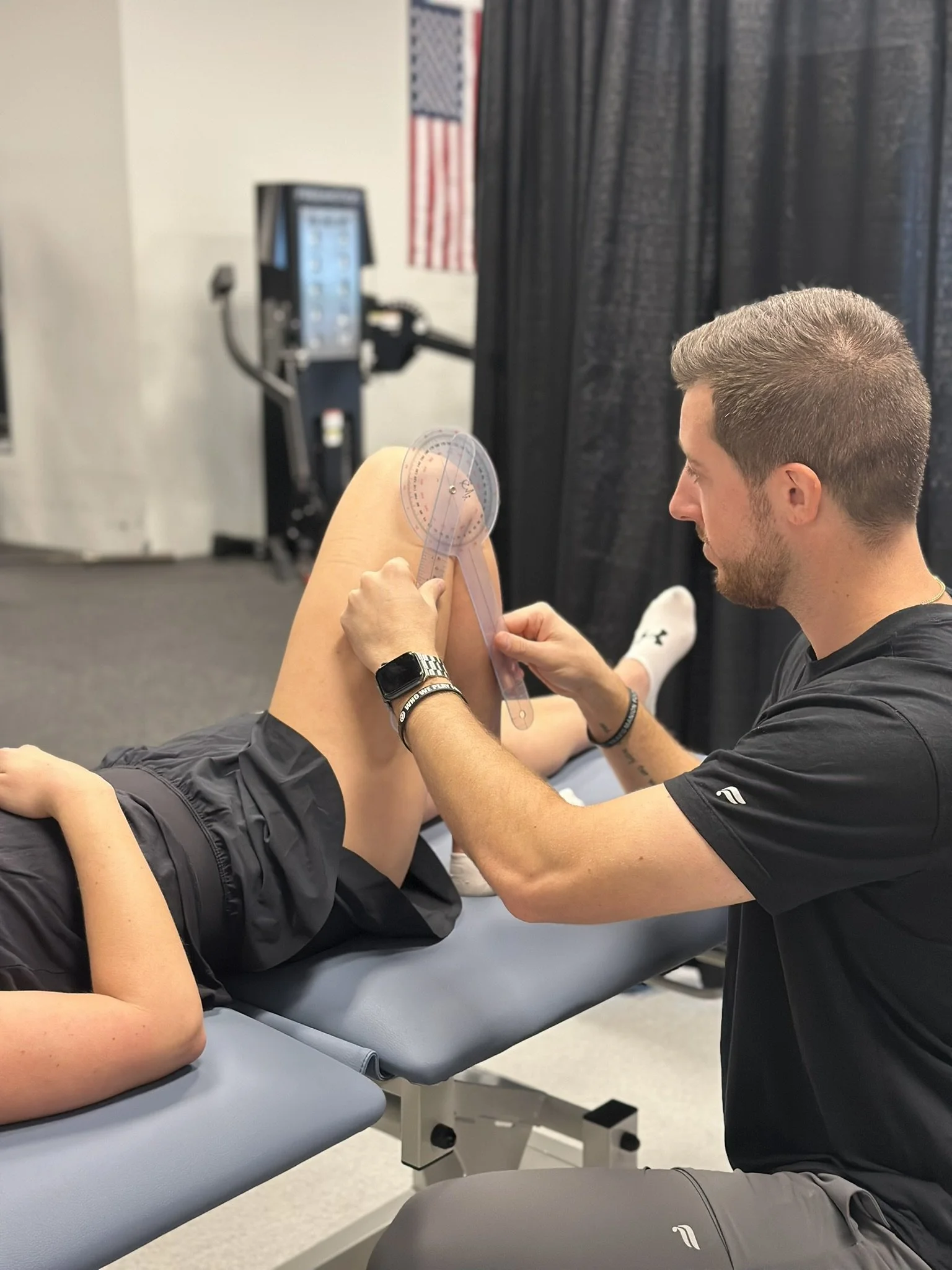 A sports physical therapist is measuring a patient's knee range of motion with a goniometer during a physical therapy evaluation, while the patient lies on an examination table.
