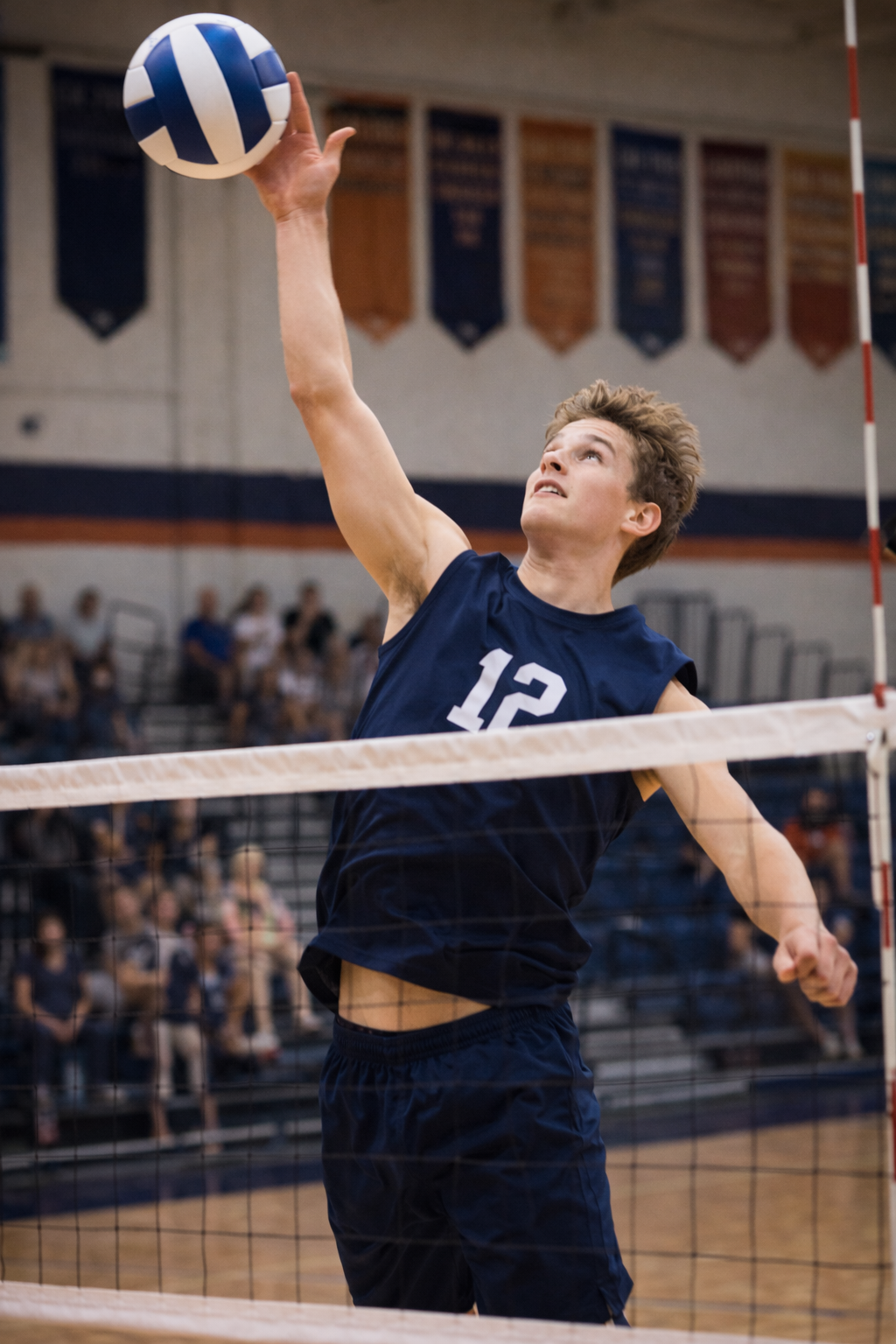A young male volleyball player wearing a navy jersey with the number 12 is jumping at the net to hit the volleyball in a gymnasium.