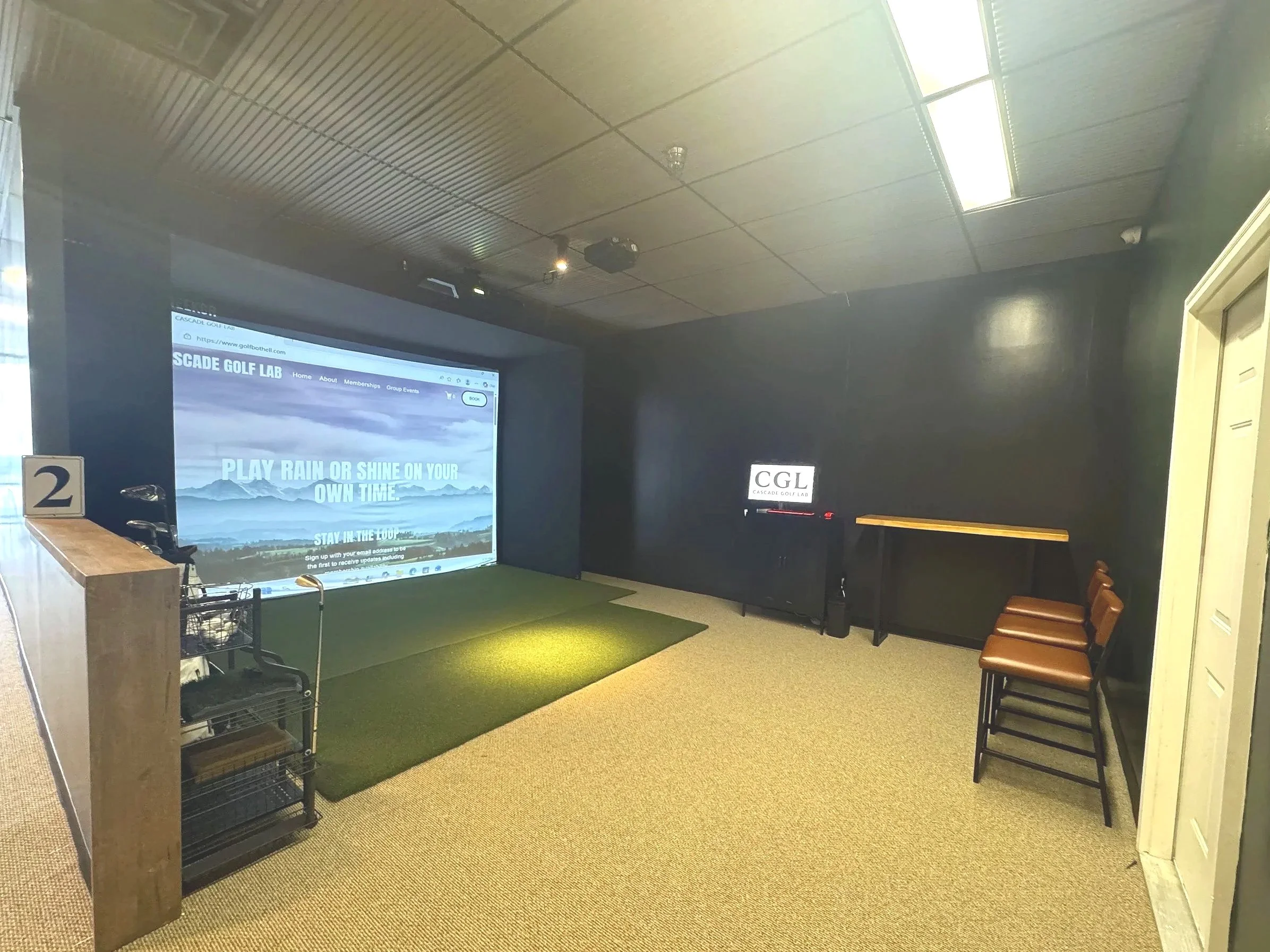 Cascade Golf Lab Bay 2- Indoor golf simulation room with a large screen displaying a golf course, chairs arranged along one wall, and a small reception counter on the left.