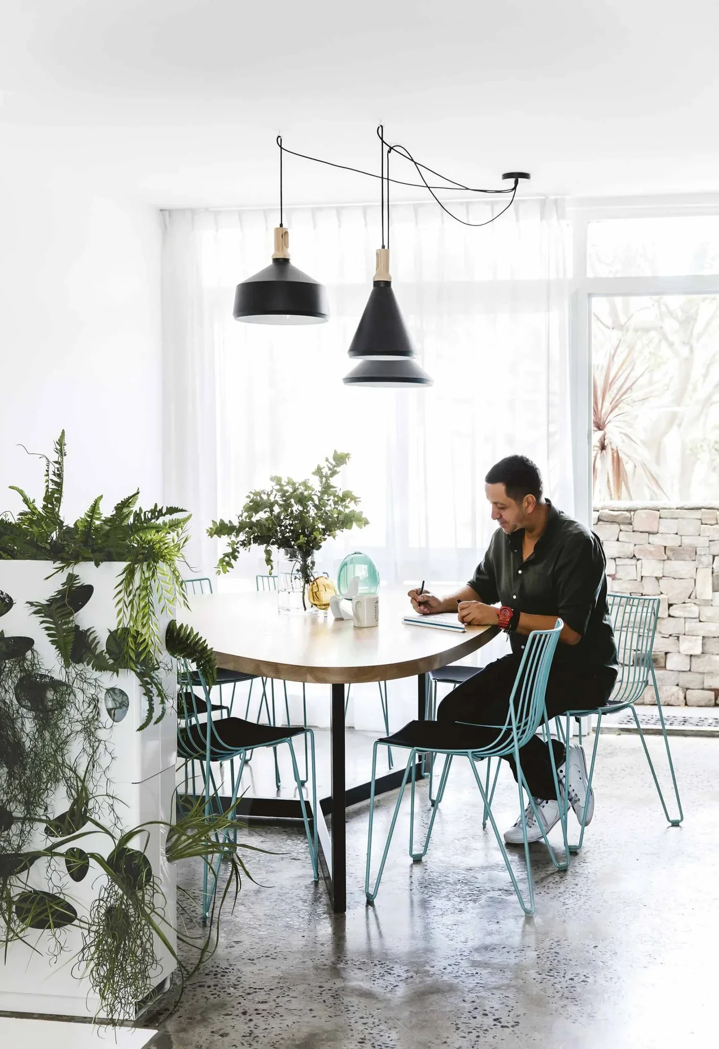 A man sitting at a dining table with plants, notebooks, and decorative objects in a brightly lit room with large windows and black pendant lights.