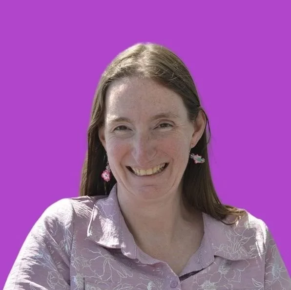 A woman with long brown hair smiling, wearing pink floral earrings and a light purple shirt with floral patterns, standing against a vibrant purple background.
