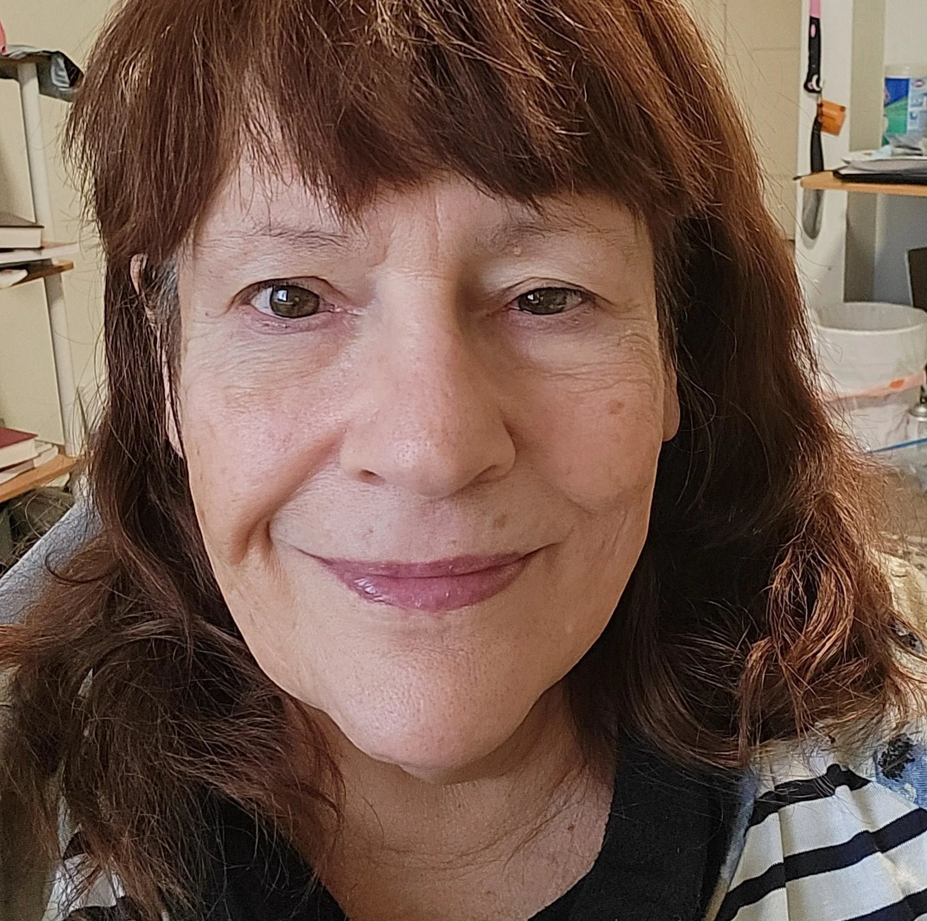 Close-up of a woman with auburn hair and a gentle smile, wearing a black top and a striped scarf, in a room with bookshelves and various objects in the background.