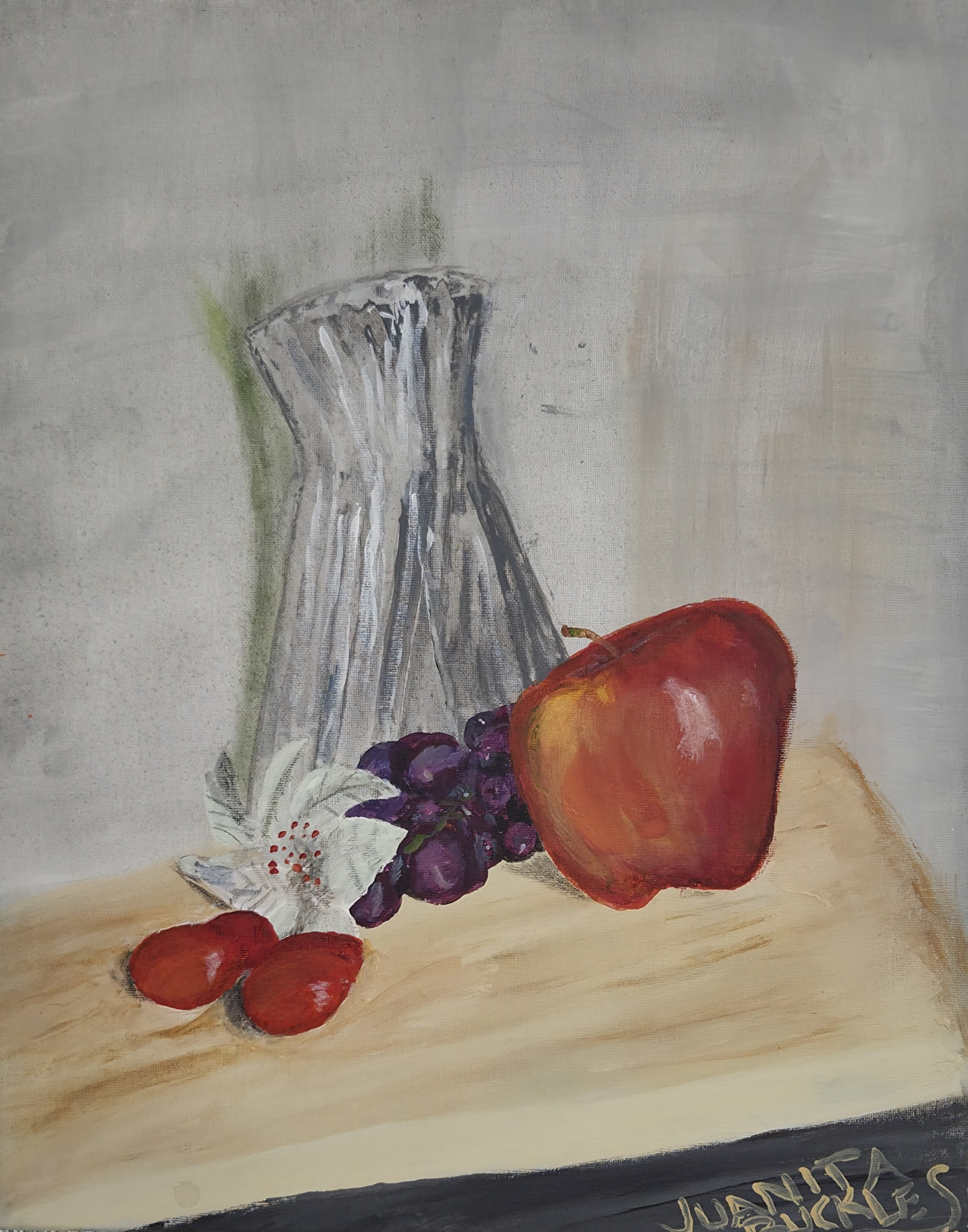 A still life painting of a glass vase, an apple, bunch of grapes, strawberry, and a white flower on a light-colored surface with a dark border at the bottom and the artist's signature.